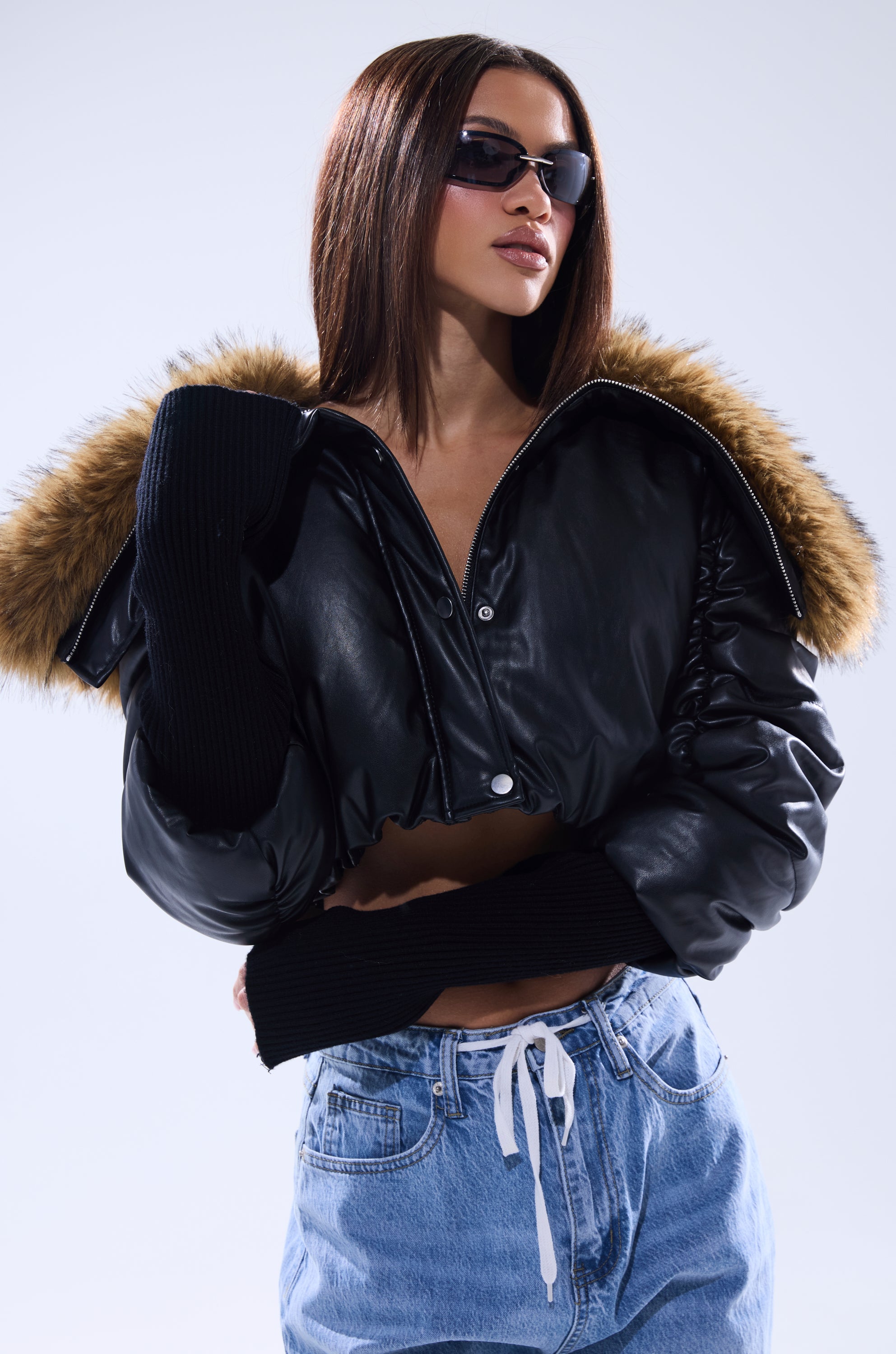 A woman confidently poses against a plain light background wearing the KICKBACK FUR TRIM BOMBER, sunglasses, and blue drawstring jeans. 2