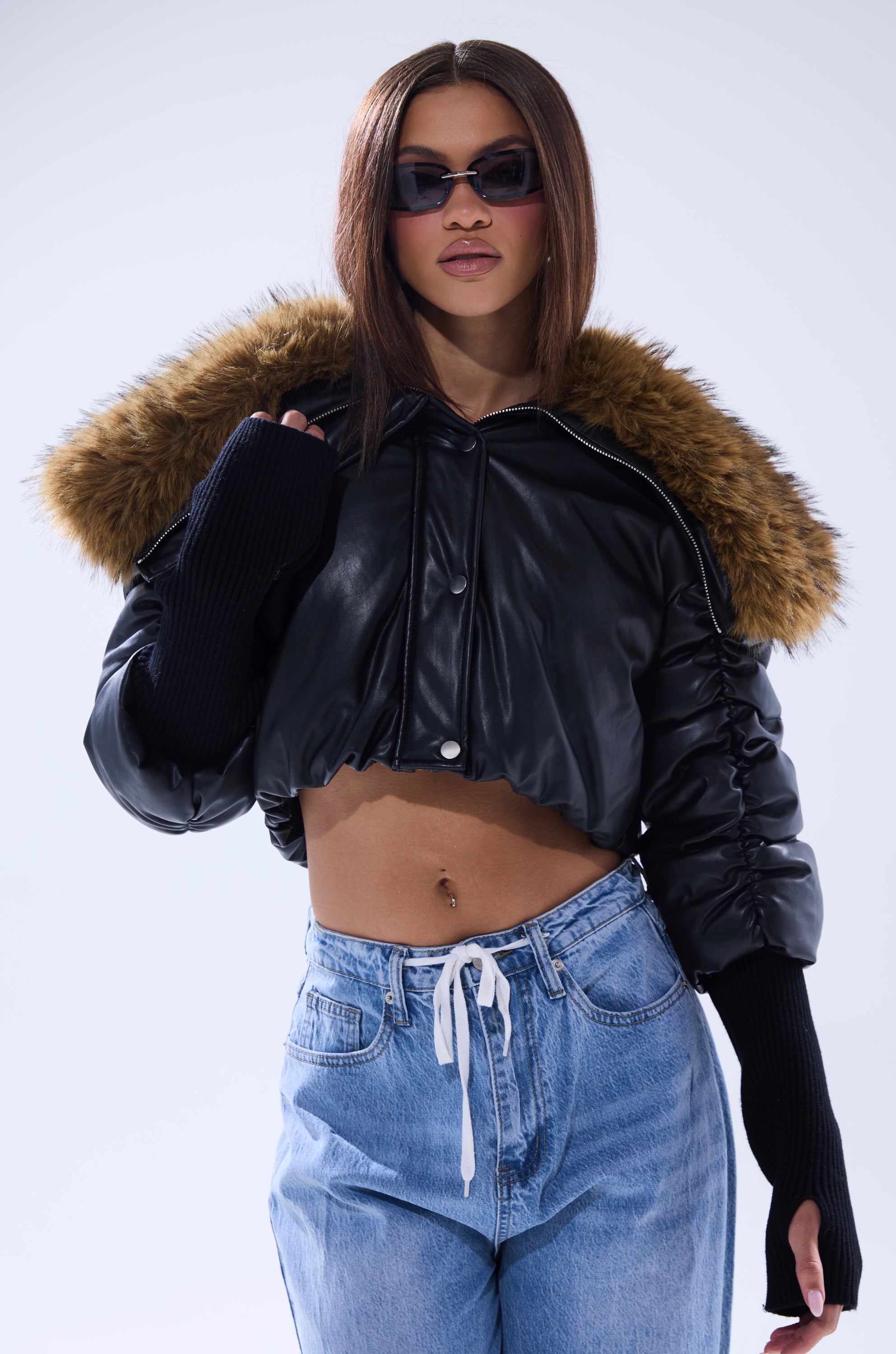 A woman stands against a plain light background wearing sunglasses, the KICKBACK FUR TRIM BOMBER with a fur-trimmed hood and long black sleeves, paired with light blue drawstring jeans. 4