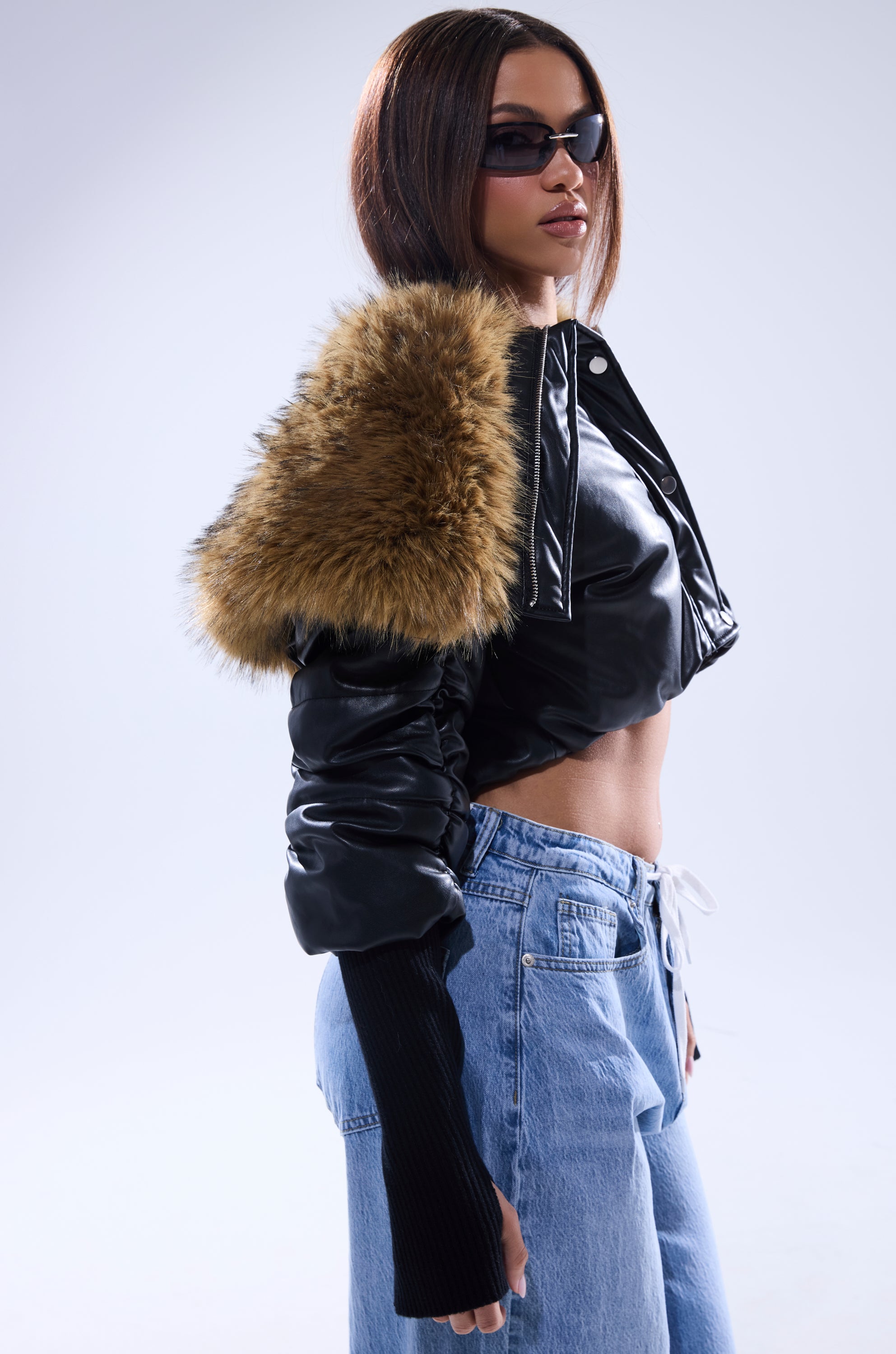 A woman stands confidently in the KICKBACK FUR TRIM BOMBER, a black cropped puffy jacket with brown faux fur shoulders, paired with light blue drawstring jeans and sunglasses against a plain light background. 5