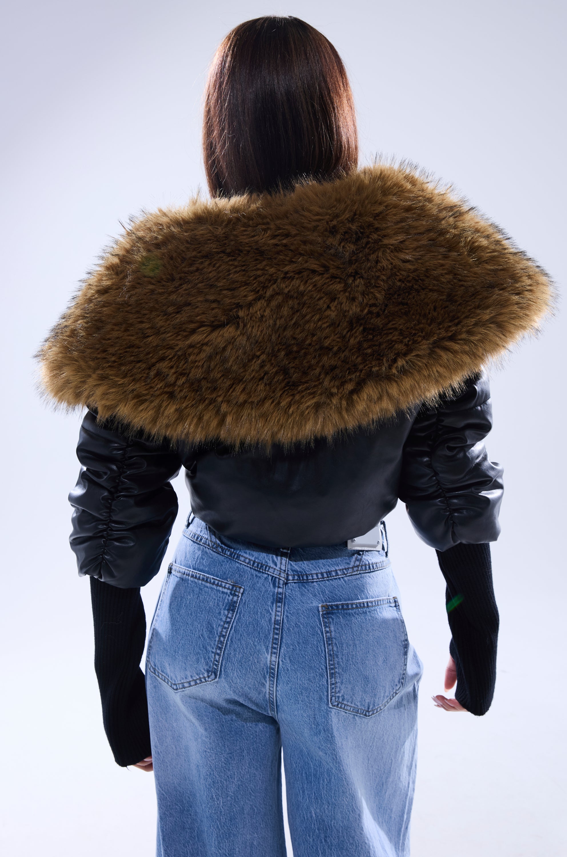 A person with straight brown hair is seen from behind wearing the KICKBACK FUR TRIM BOMBER, which features a black design with a large brown collar, paired with blue jeans against a plain light background. 6