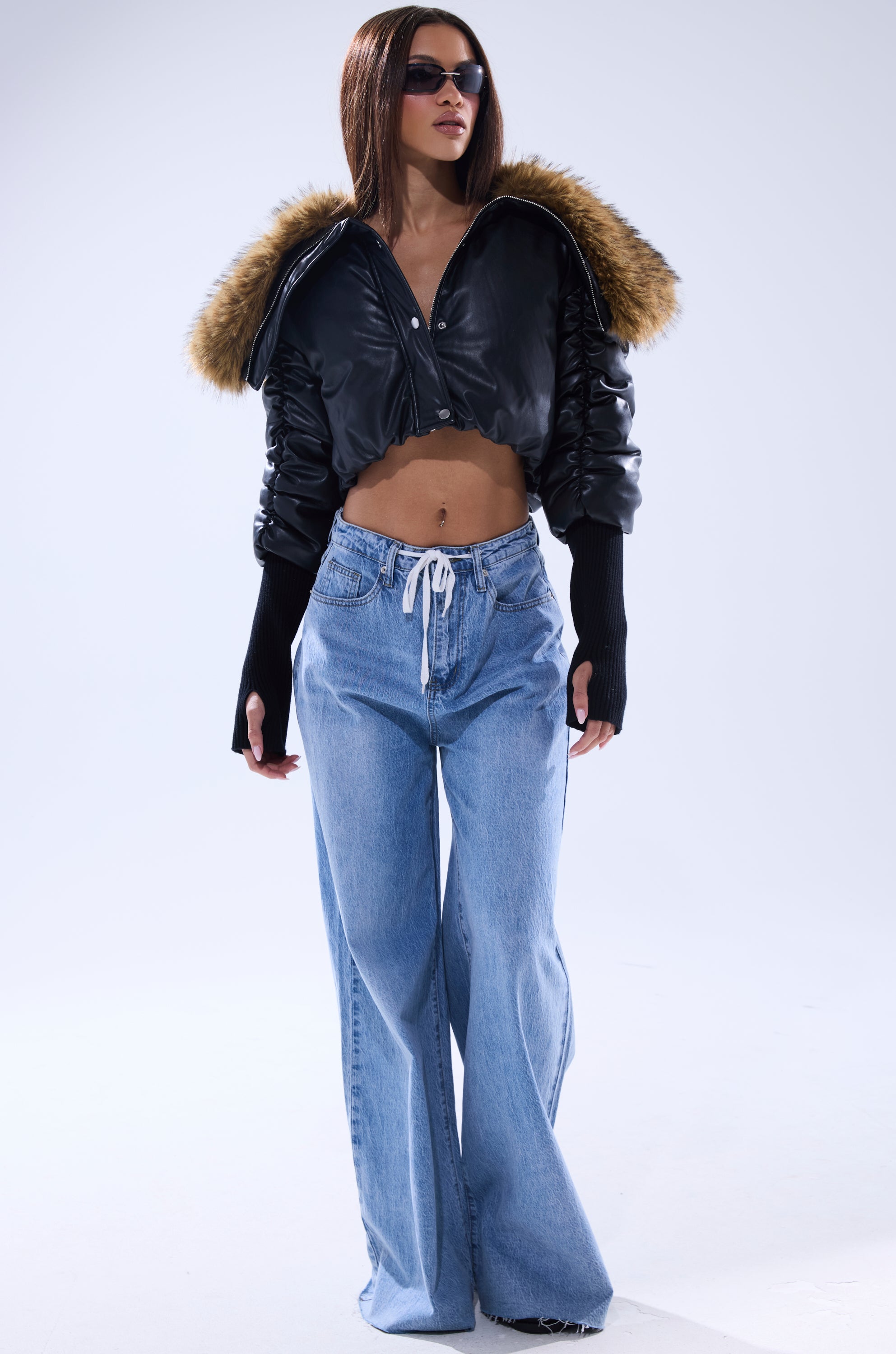 A woman with straight brown hair and sunglasses wears the KICKBACK FUR TRIM BOMBER, a cropped puffy jacket with a fur-trimmed hood, wide-leg blue jeans, and long black sleeves against a plain light background. 8