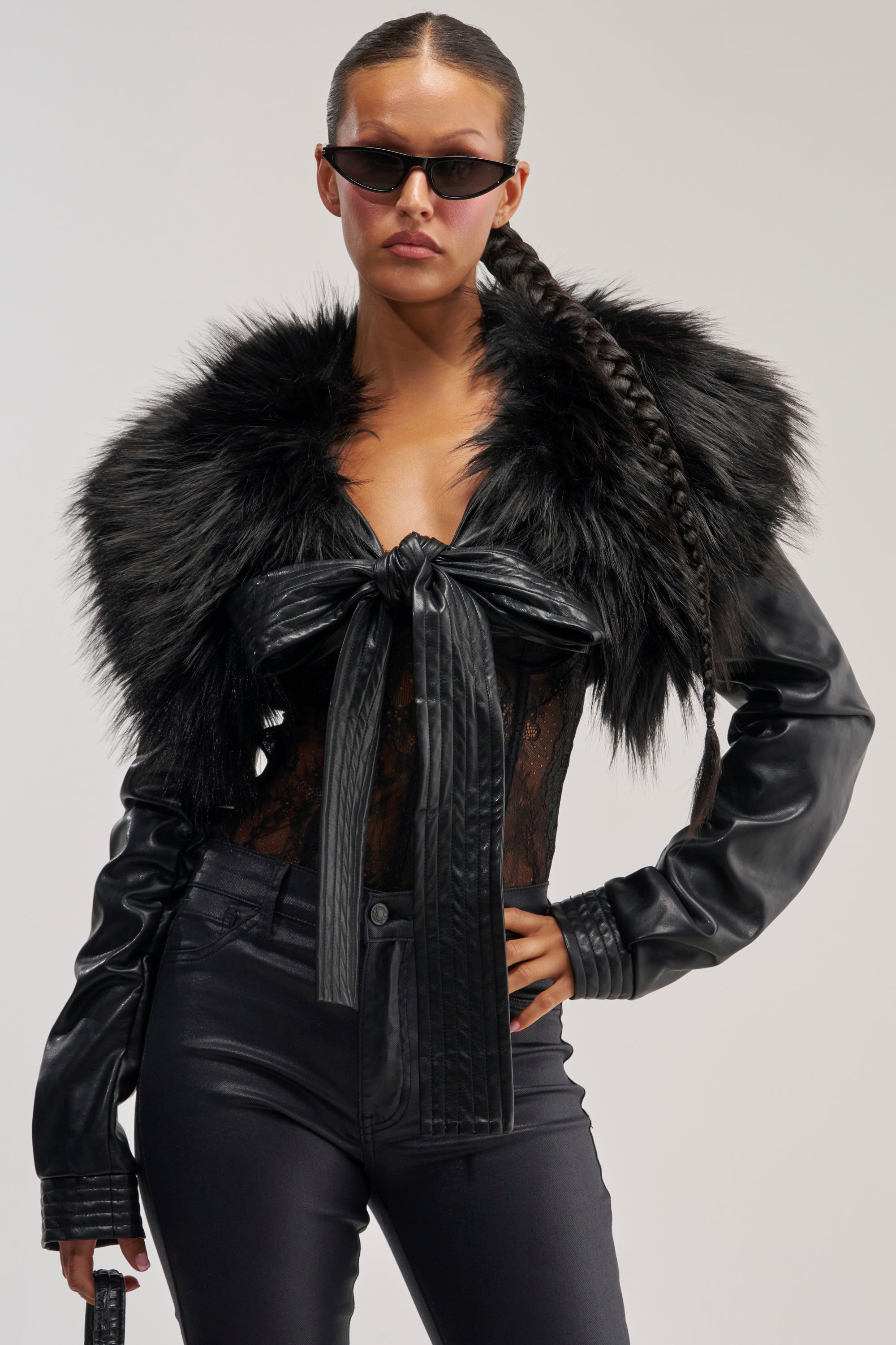 A woman in sunglasses wears the SPEND IT ALL FUR TRIM BOLERO IN BLACK over a black leather jacket with a large bow, paired with a black lace top and black pants. With her long braid and confident stance against a plain background, she perfectly embodies chic winter layering.