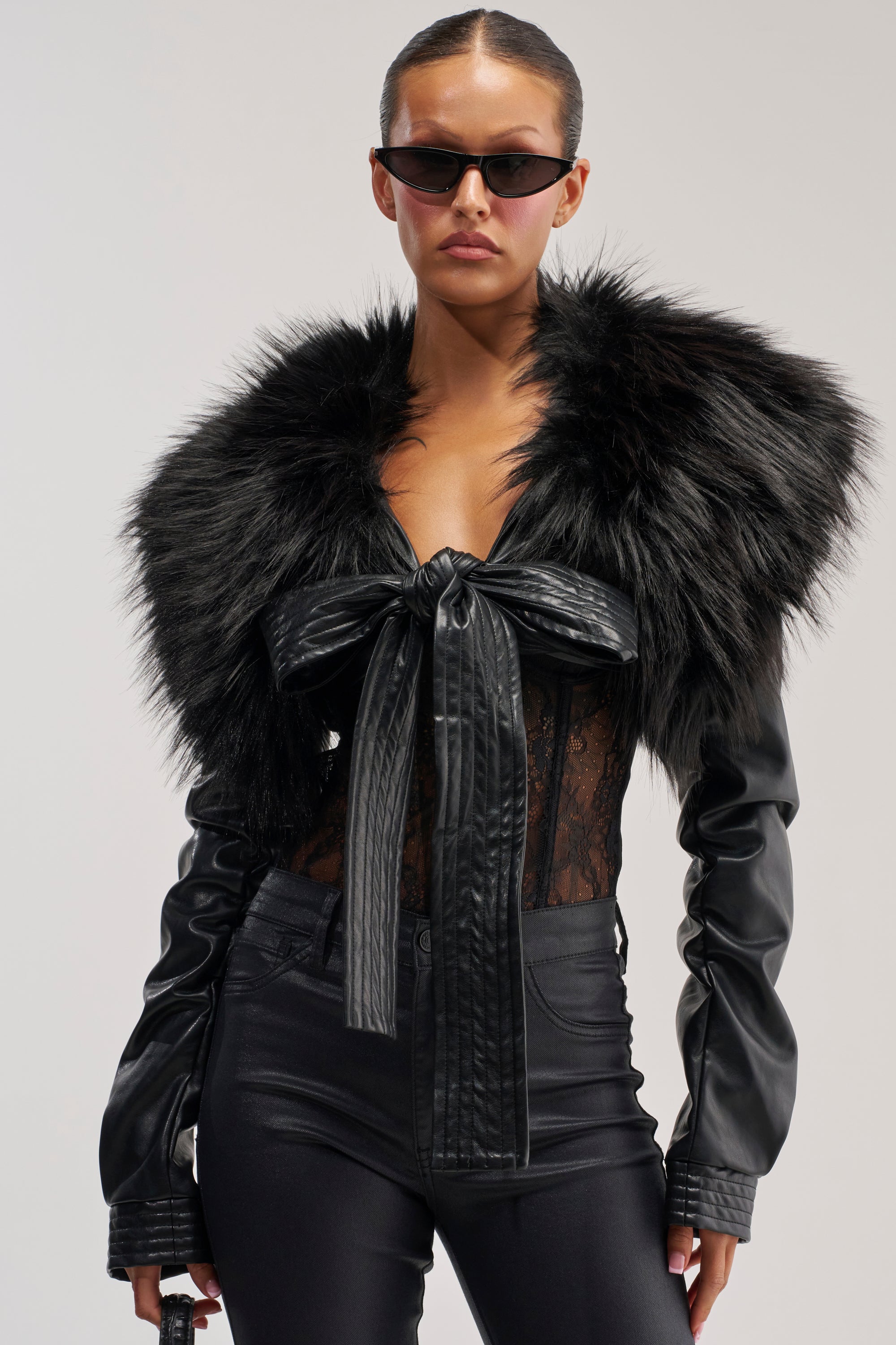 Woman wearing sunglasses, the SPEND IT ALL FUR TRIM BOLERO IN BLACK featuring a large leather bow, styled with a sheer lace top and black leather pants, posing against a plain light background—perfect for chic winter layering.