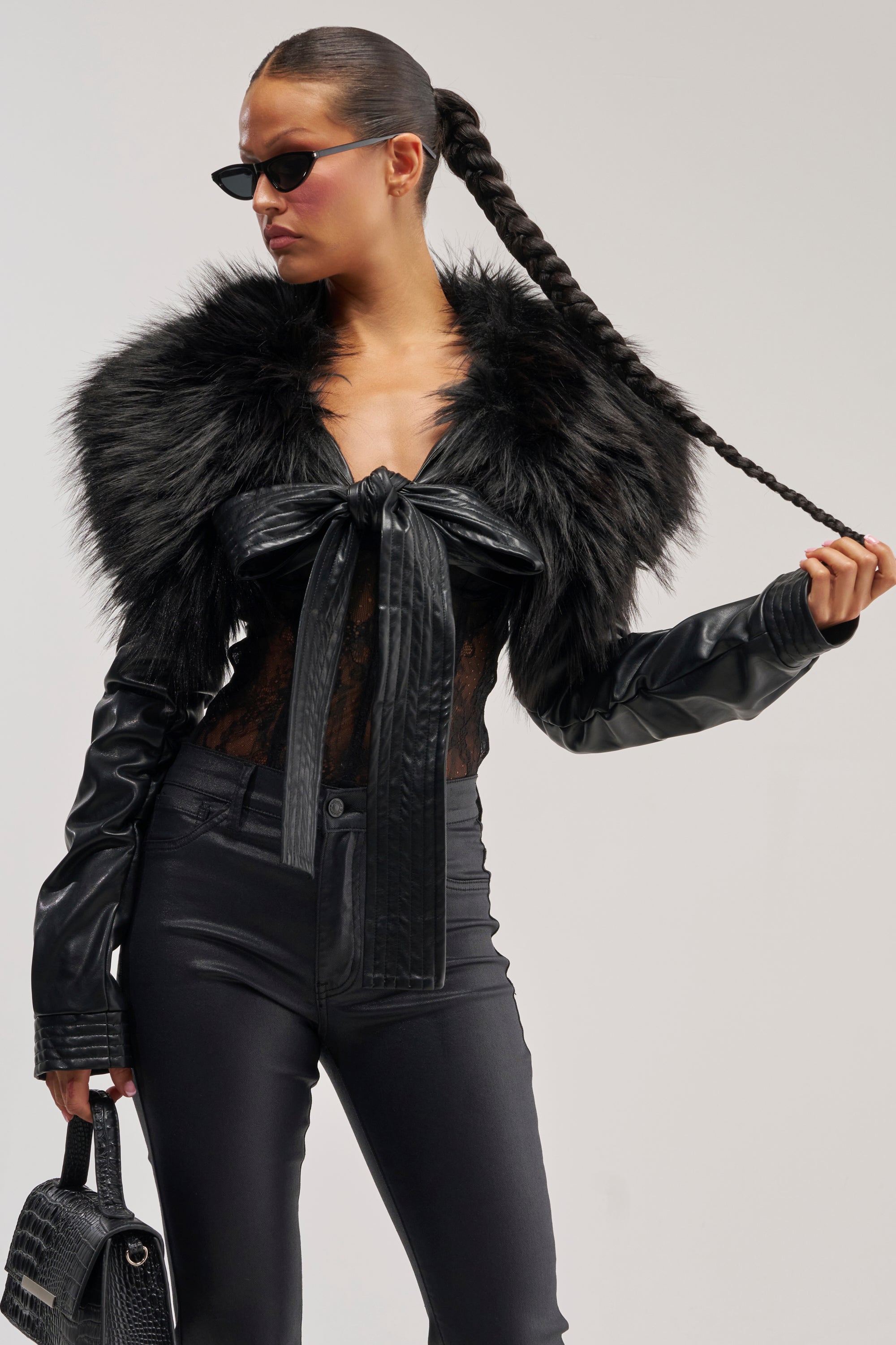 A woman in black sunglasses, leather pants, and the SPEND IT ALL FUR TRIM BOLERO IN BLACK layered over a lace top holds her long braided ponytail and a handbag as she poses confidently against a plain background.