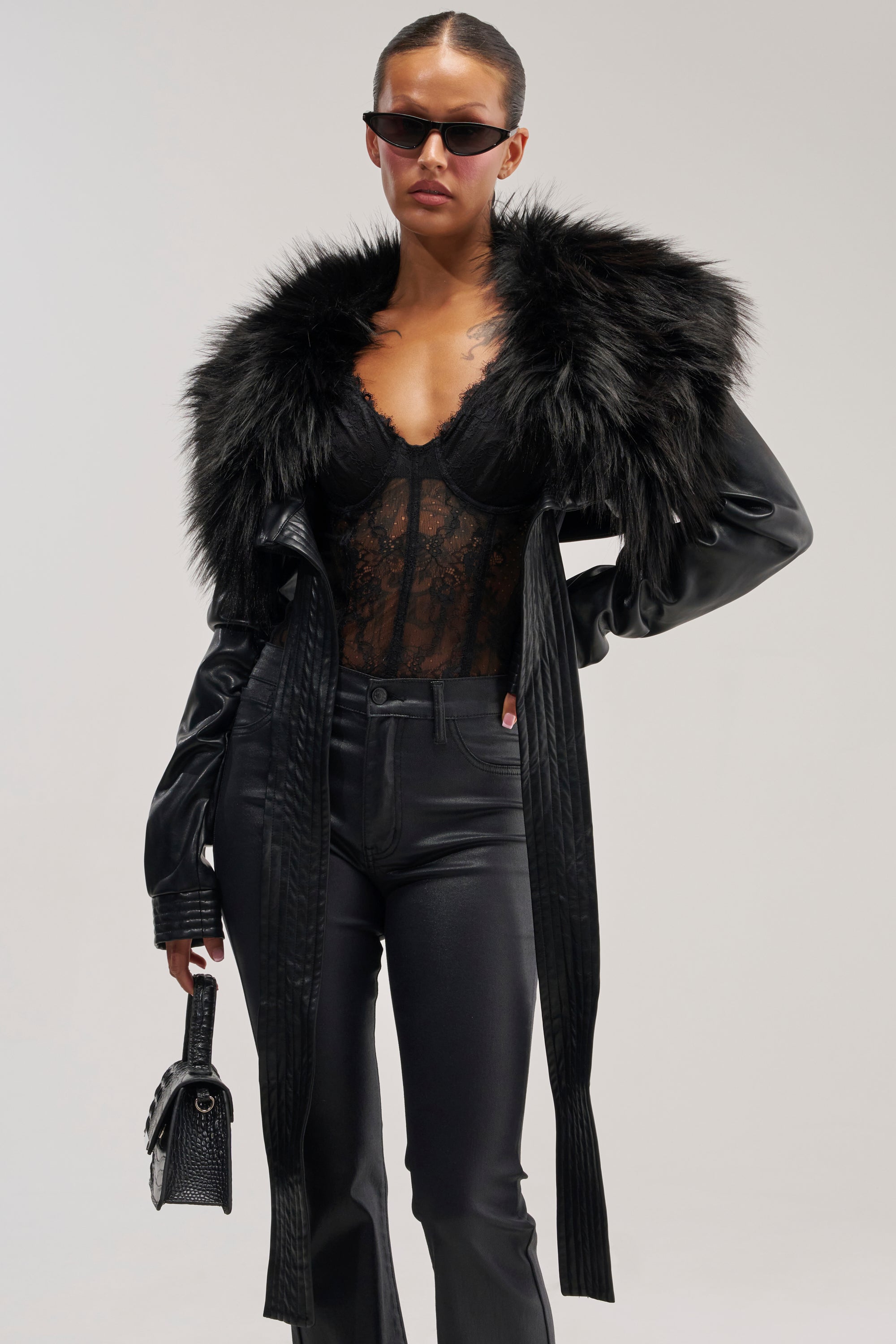 Wearing the SPEND IT ALL FUR TRIM BOLERO IN BLACK, a woman in sunglasses pairs its luxurious fur collar with a black lace top and black pants—an ideal look for winter layering. She carries a small black handbag and stands against a simple light background.