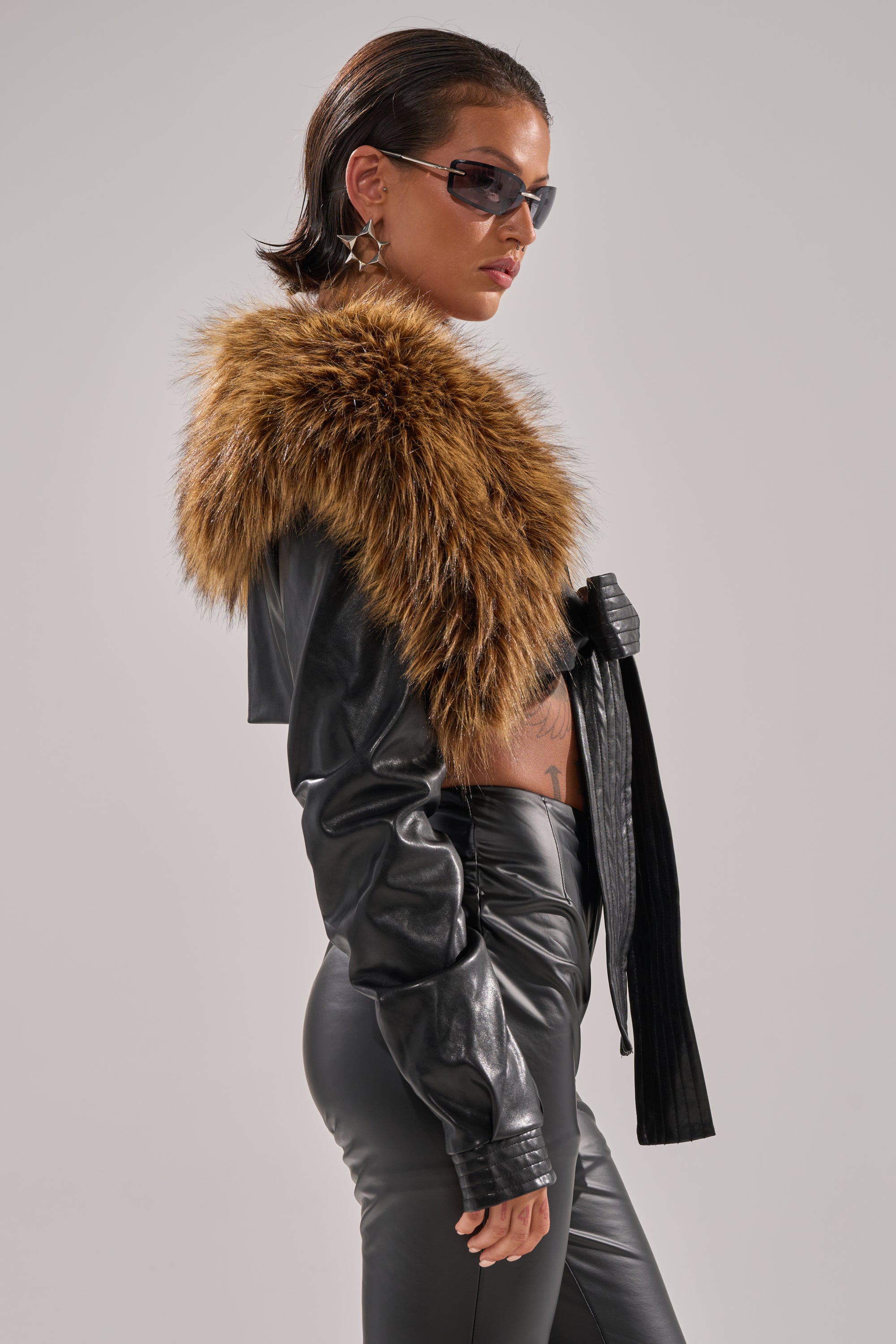A person in the SPEND IT ALL FUR TRIM BOLERO with a large brown collar, black leather pants, and sunglasses stands in profile against a plain light background.