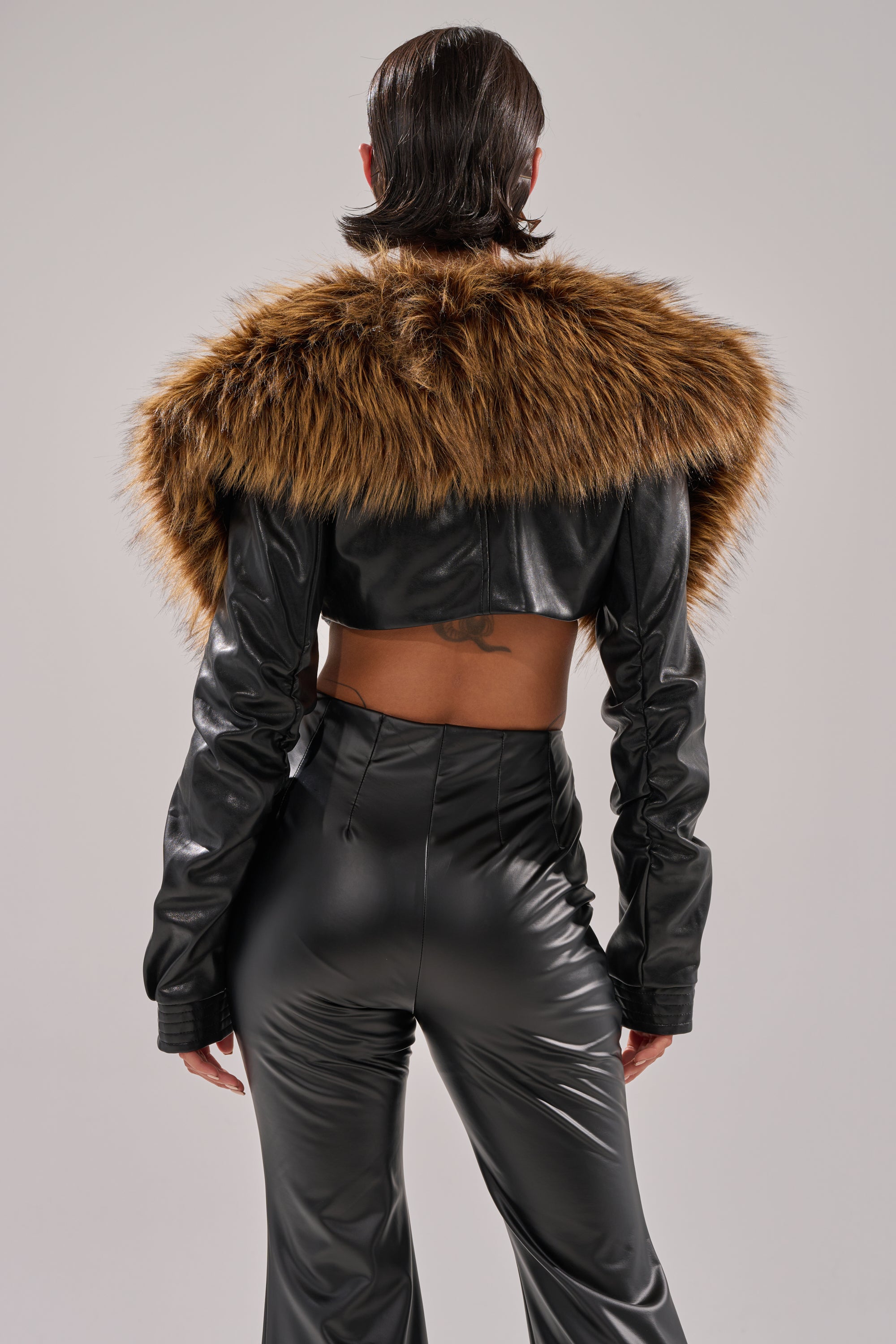 A person wears shiny black faux leather pants and the SPEND IT ALL FUR TRIM BOLERO with a large brown faux fur collar, photographed from the back against a plain background.