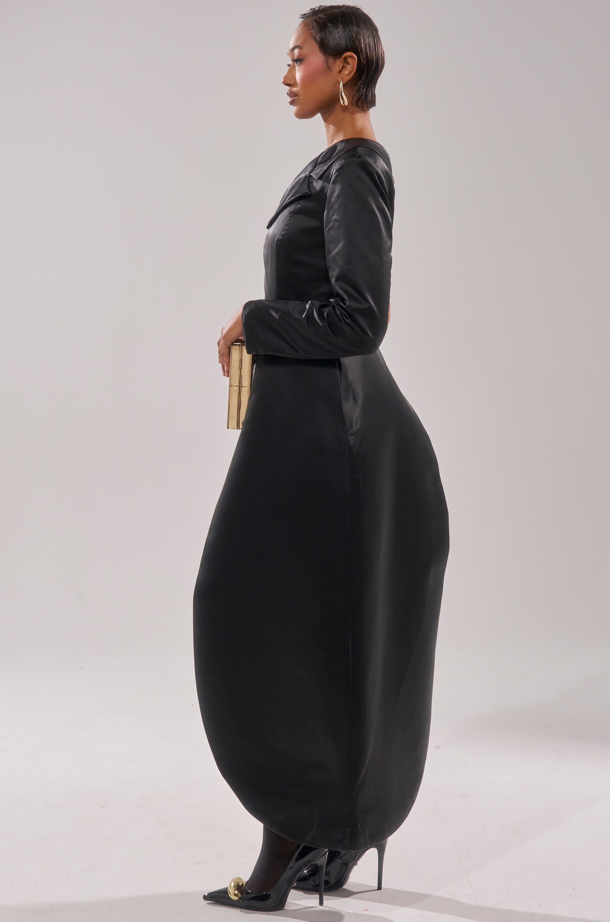 A woman stands in profile wearing the BLACK TIE AFFAIR TUXEDO TOP and a long, voluminous black skirt, paired with black heels, gold earrings, and a small gold clutch against a plain light background.