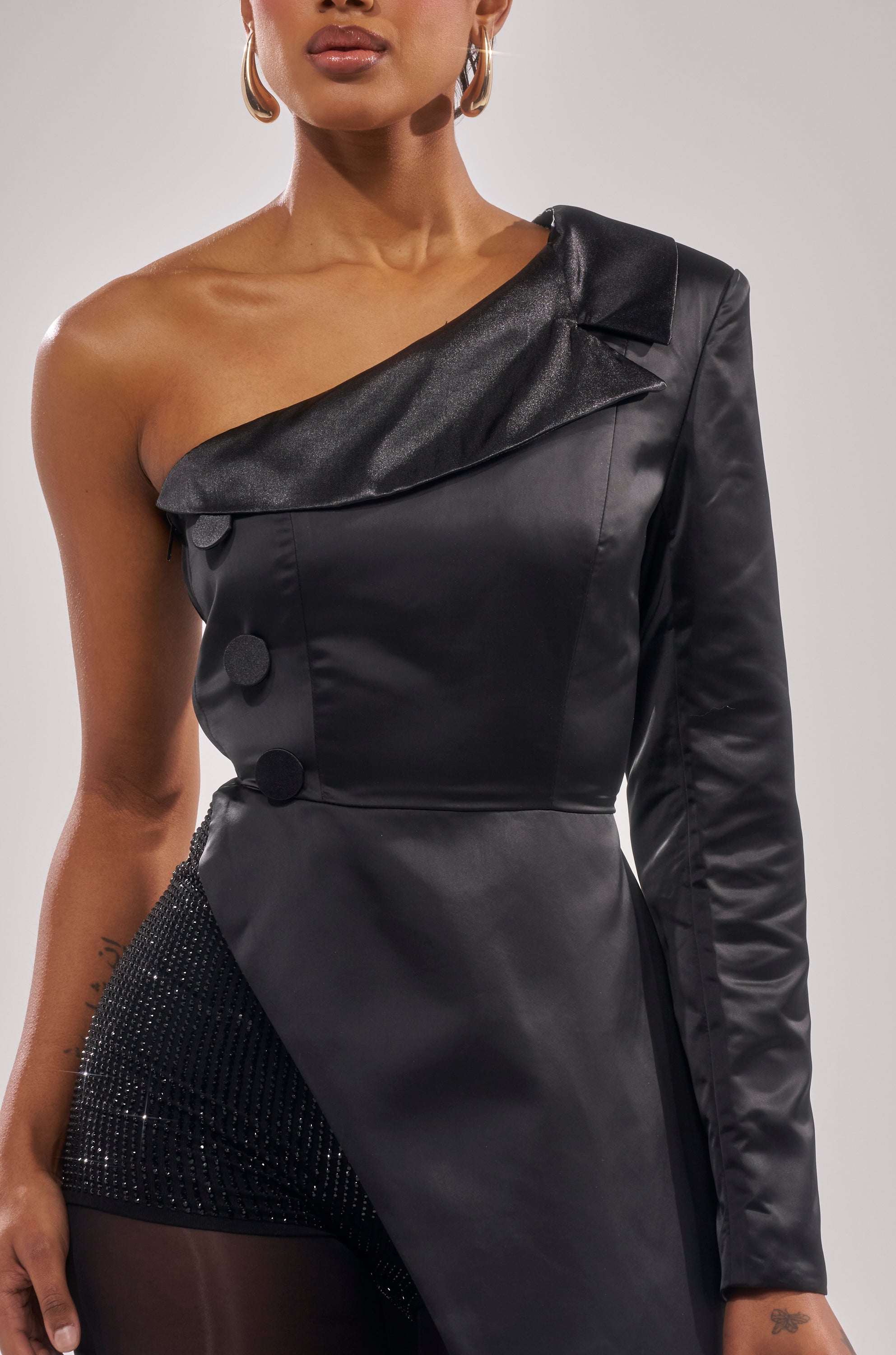 A woman wears the BLACK TIE AFFAIR TUXEDO TOP, a sleek black one-shoulder piece with an asymmetrical neckline, bold buttons, and textured details. Gold hoop earrings and a small floral wrist tattoo complete her chic look.