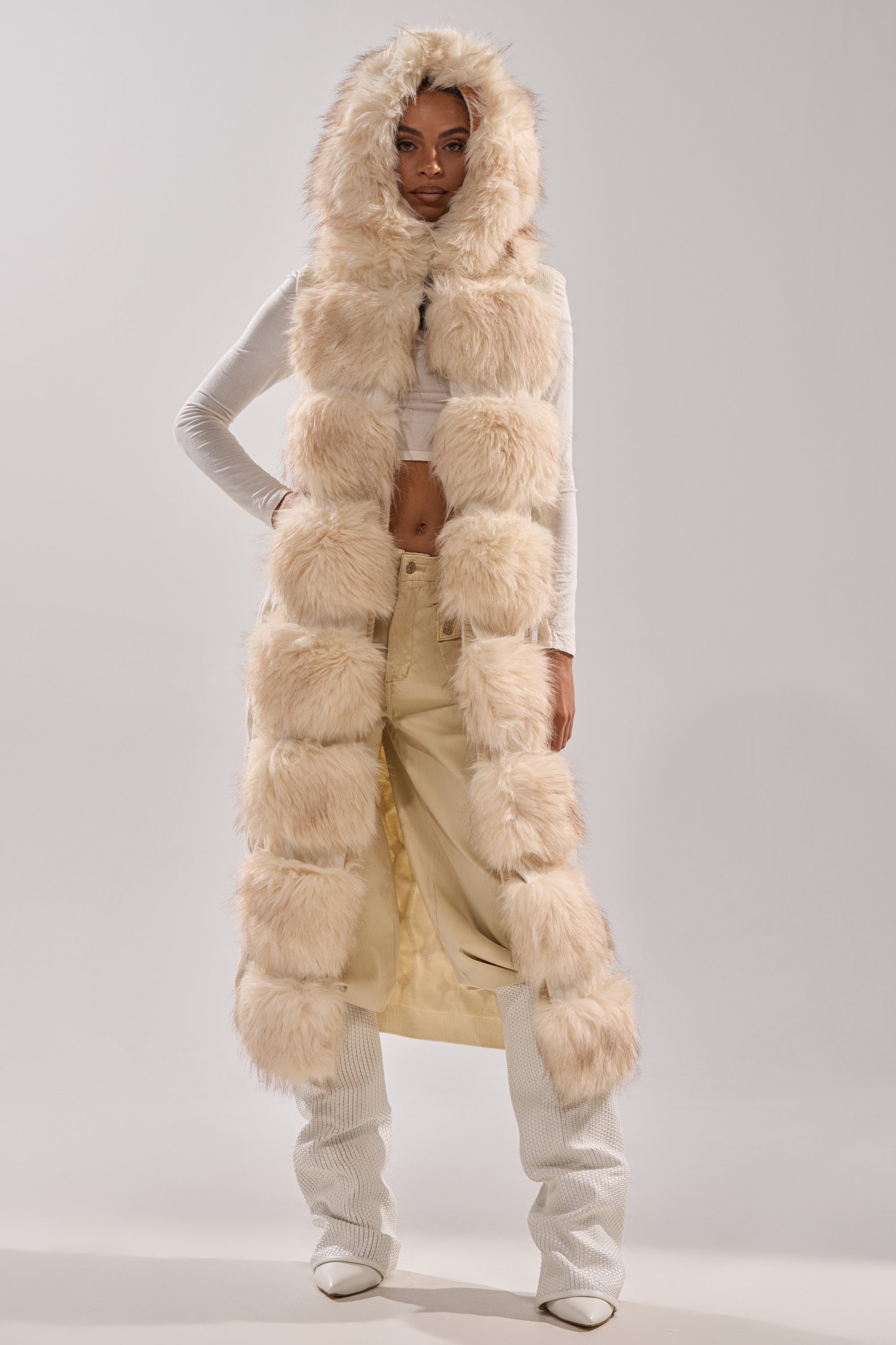 A woman models the LUXURIOUS FAUX FUR HOODED VEST in cream beige, paired with a white long-sleeve crop top, wide-leg beige pants, and white shoes against a plain light background.