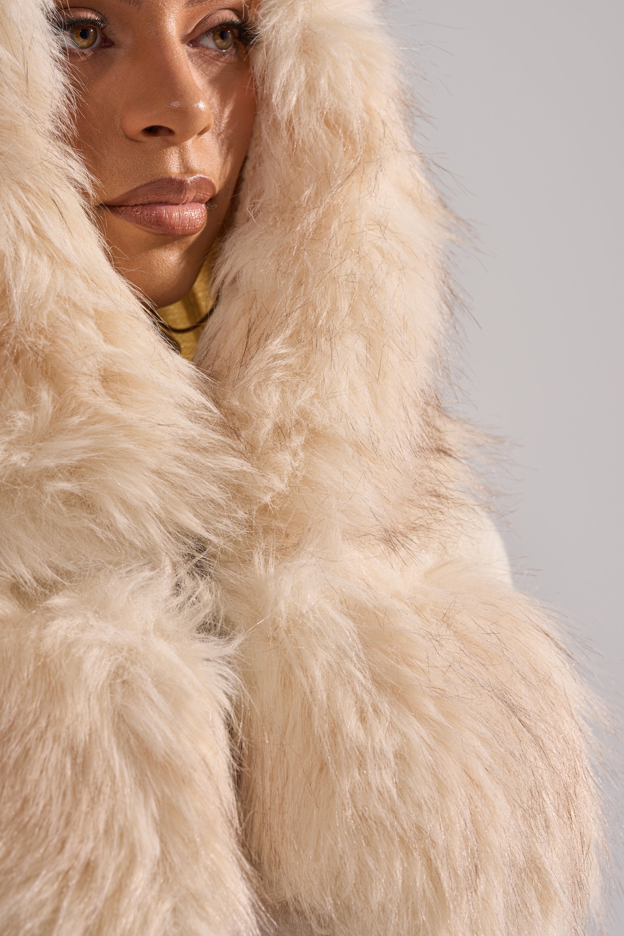 A close-up shows a person in the LUXURIOUS FAUX FUR HOODED VEST, its large cream-colored faux fur hood partially covering their face, leaving only the lips, nose, and one eye visible under soft, neutral lighting.