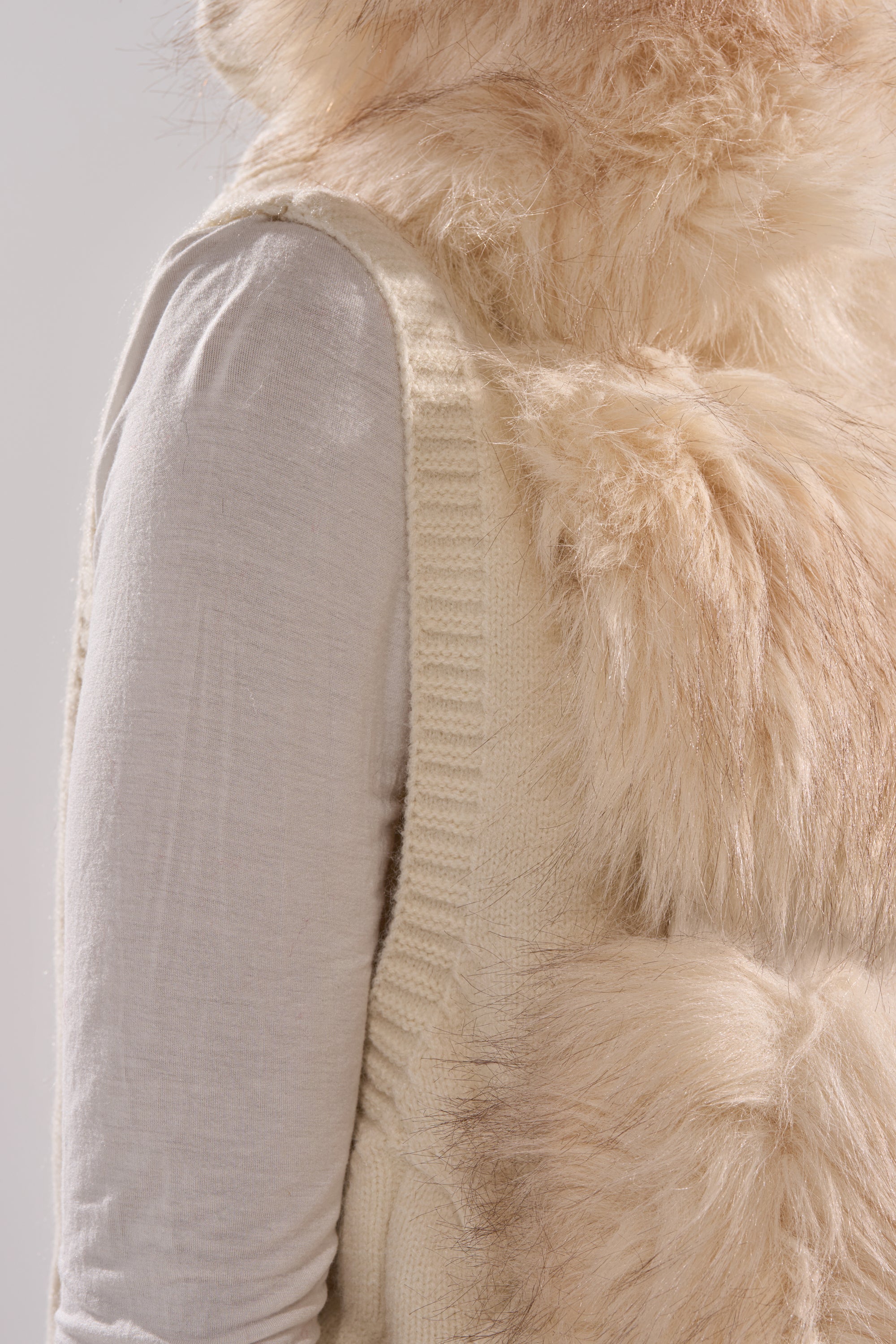 A person is shown from the side against a plain light background wearing the LUXURIOUS FAUX FUR HOODED VEST, a fluffy light beige layer styled over a cream long-sleeve shirt.