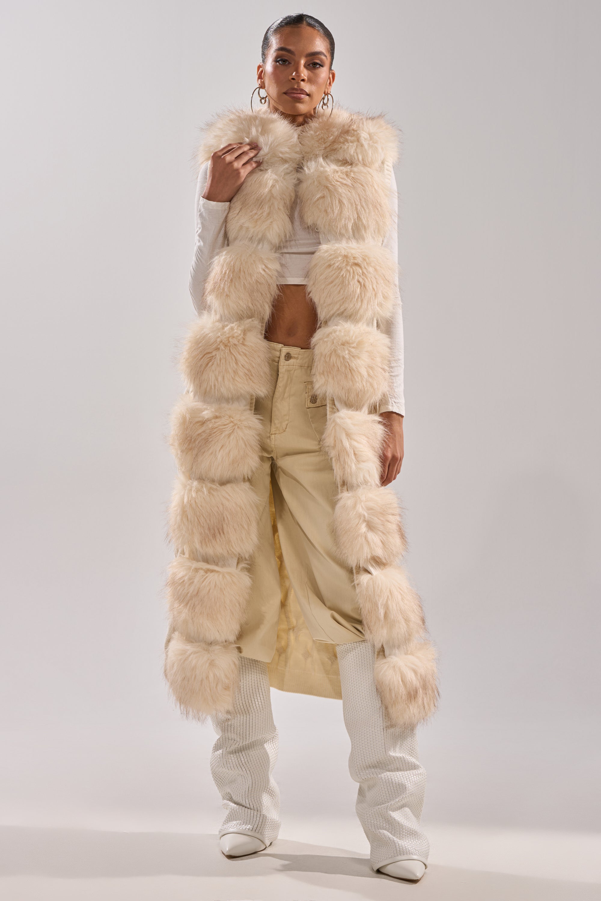 A person faces forward, wearing the LUXURIOUS FAUX FUR HOODED VEST over a white cropped top and beige pants with white textured boots, set against a plain light gray background.