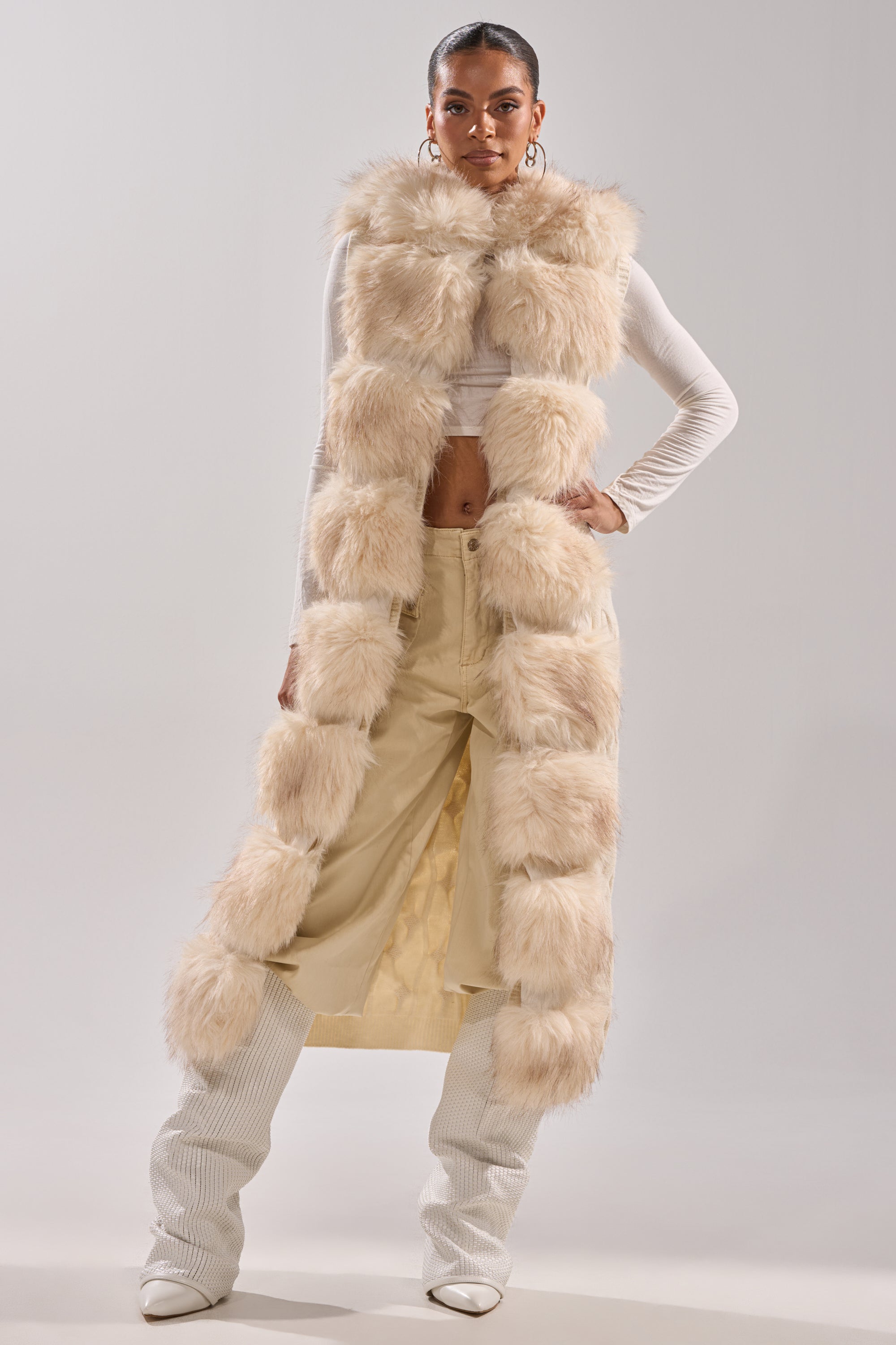 A woman poses confidently in the LUXURIOUS FAUX FUR HOODED VEST over a white crop top and beige high-waisted pants, paired with white heeled boots and large hoop earrings, standing against a plain background.