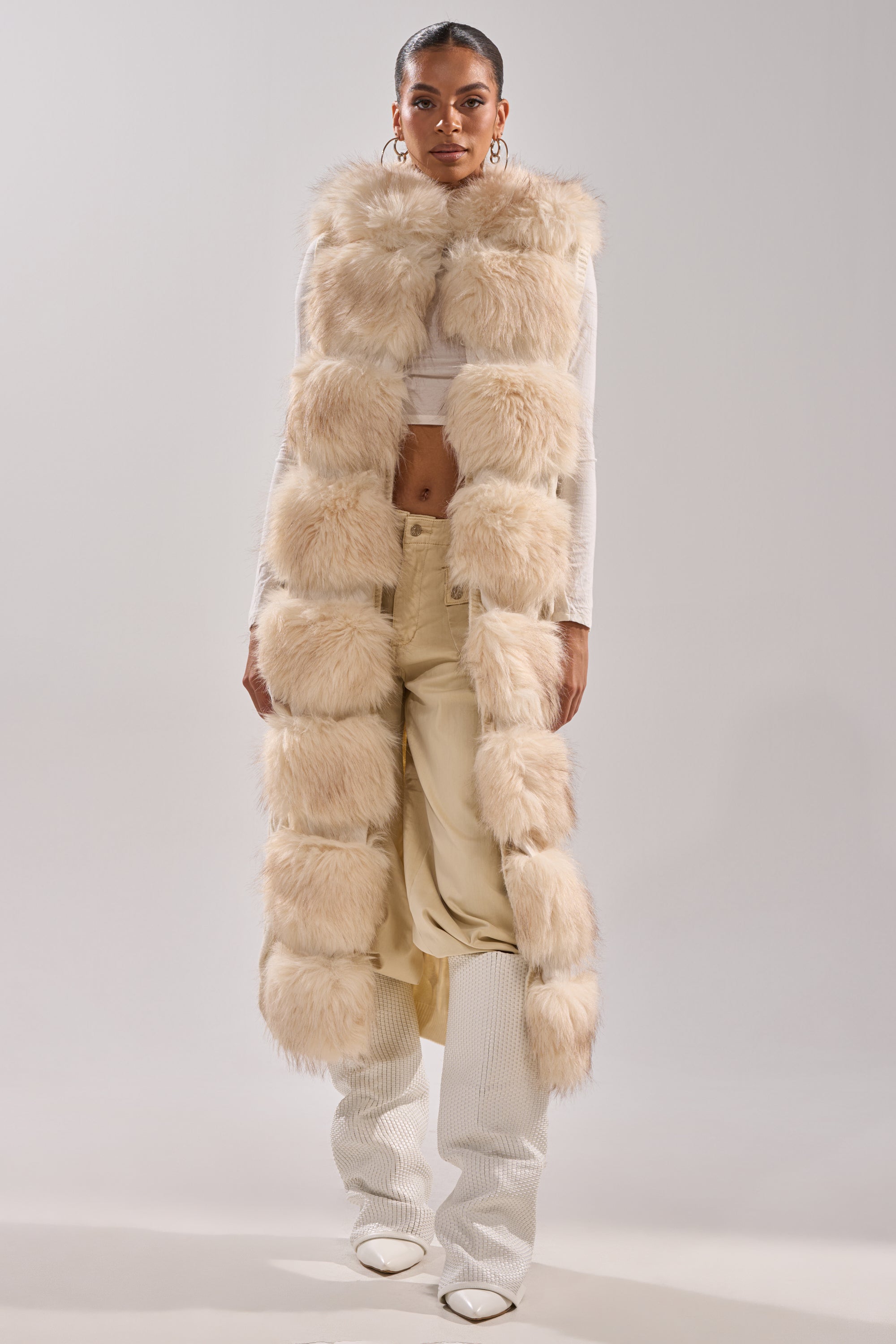 A woman models the LUXURIOUS FAUX FUR HOODED VEST over a white crop top and cream pants with ribbed leg warmers and white pointed shoes, posing against a plain light background.