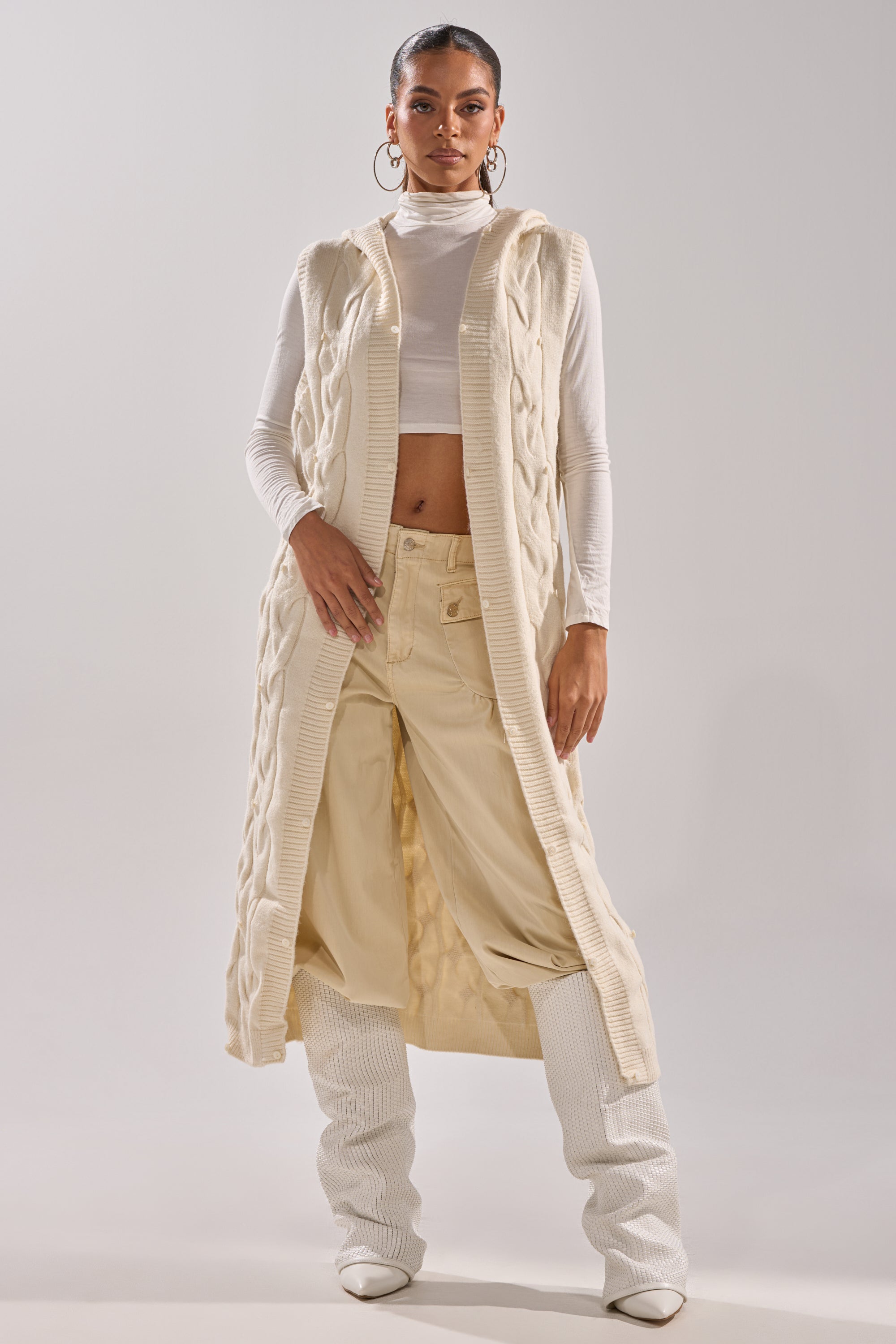 A woman poses confidently in the LUXURIOUS FAUX FUR HOODED VEST layered over a white turtleneck crop top and loose cream pants with ribbed white boots, set against a plain light background.
