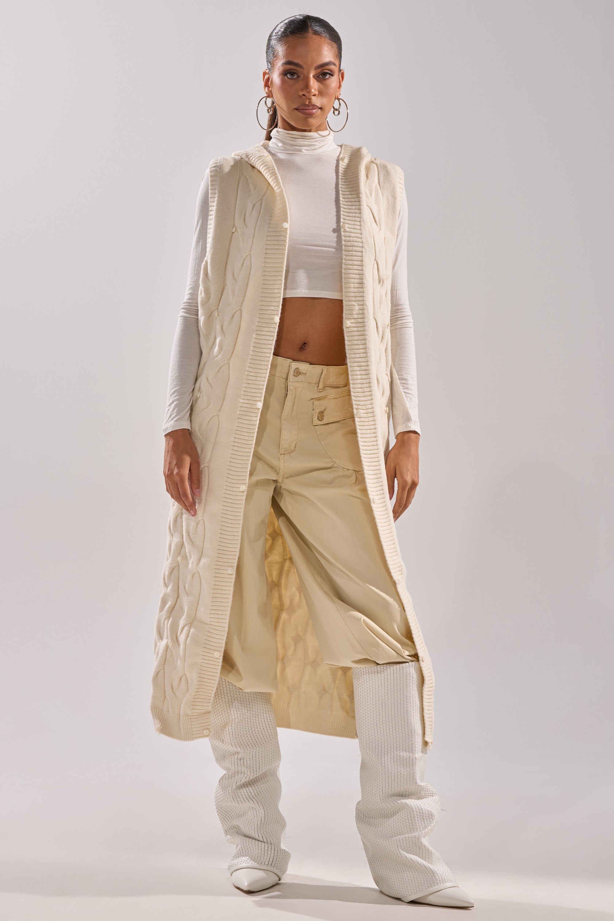 A woman faces forward in a LUXURIOUS FAUX FUR HOODED VEST over a white turtleneck, cream wide-leg pants, and white knee-high textured boots. She accessorizes with hoop earrings and has her hair pulled back.