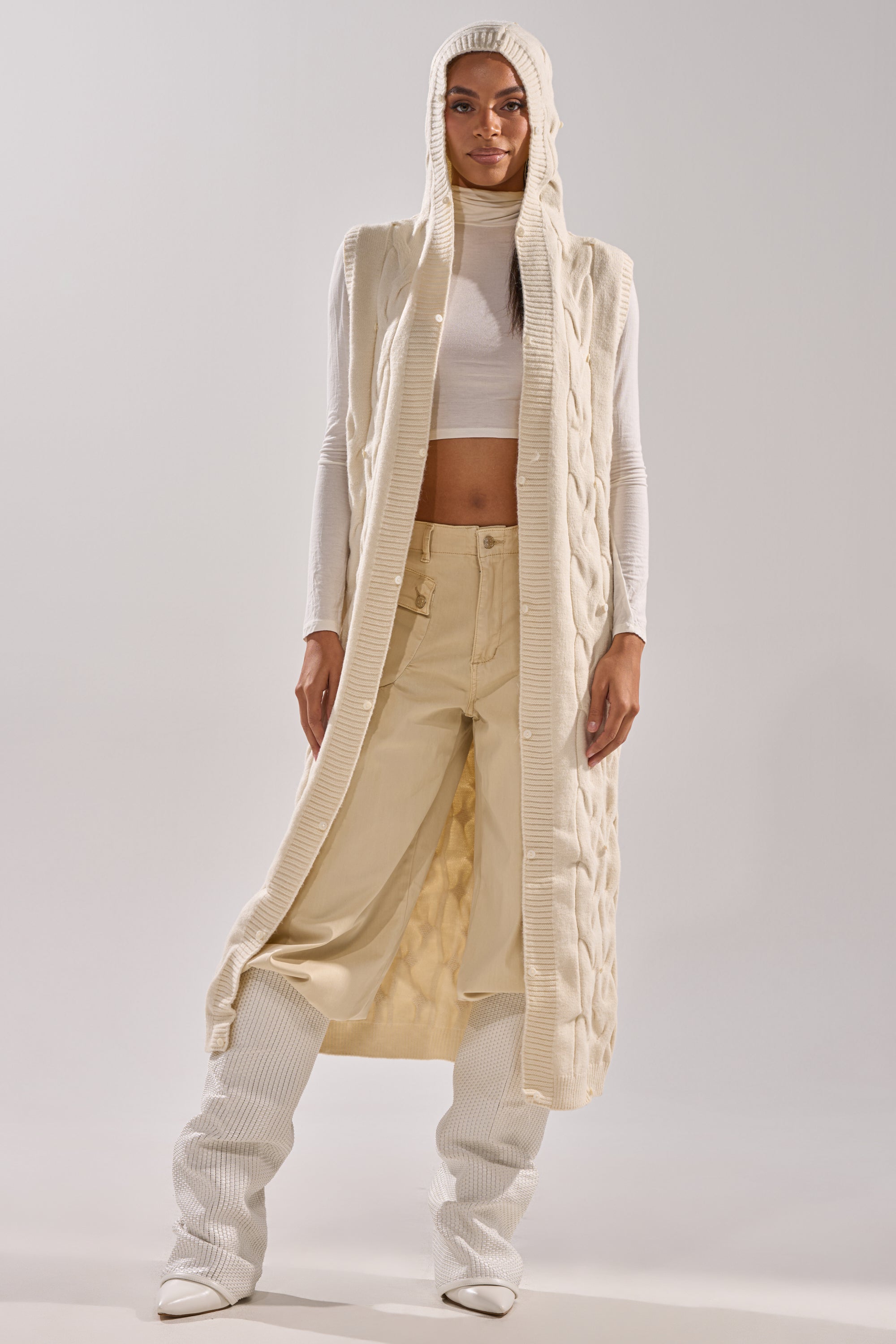 A woman stands facing forward in the LUXURIOUS FAUX FUR HOODED VEST, styled with a white cropped turtleneck, cream pants, and white heeled boots against a plain background.