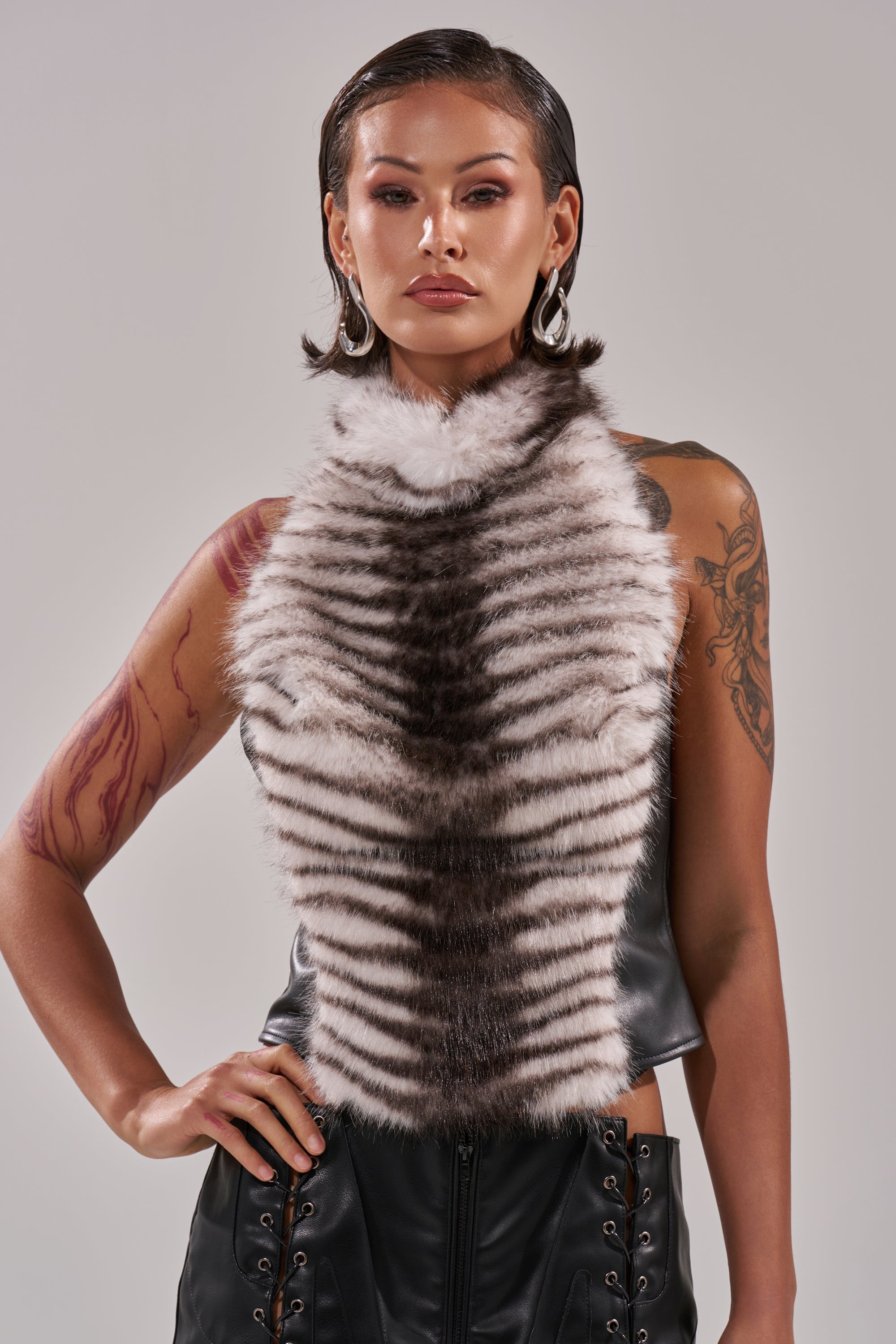 A confident woman with slicked-back hair wears the I'M MY OWN SPIRIT ANIMAL MOCK NECK FUR TOP, featuring a striped, furry front and cropped fit. She poses with hoop earrings, bold makeup, visible tattoos, and one hand on her hip. 0