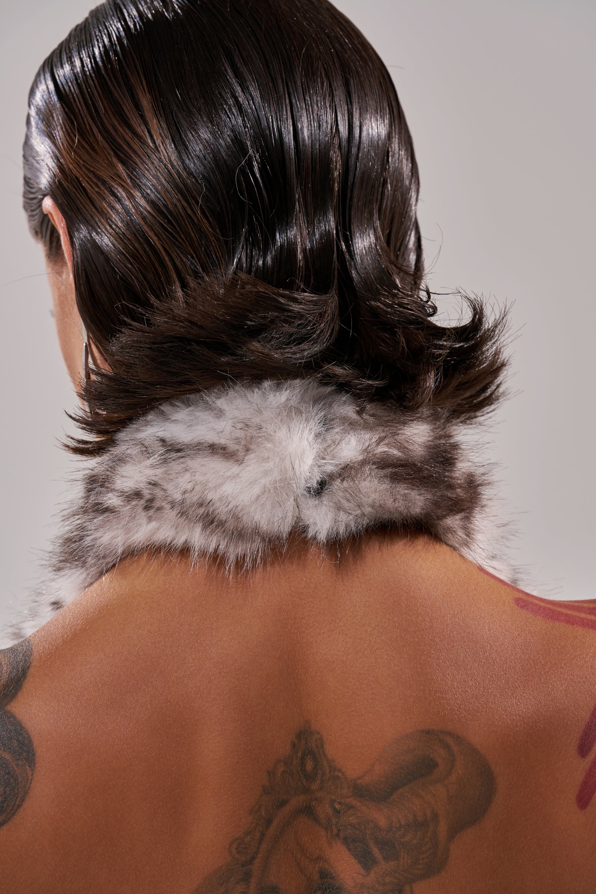 A person with wet, slicked-back dark hair is shown from behind in the I'M MY OWN SPIRIT ANIMAL MOCK NECK FUR TOP, a cropped gray fur style. Their bare back features tattoos, including a large snake on the upper back. 9
