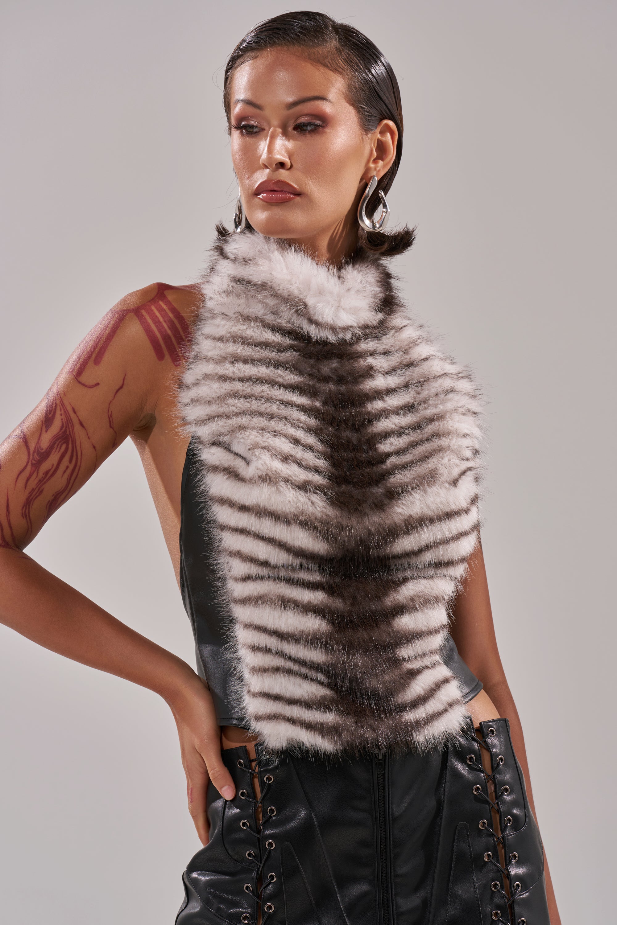 Wearing the I'M MY OWN SPIRIT ANIMAL MOCK NECK FUR TOP, a person with slicked-back hair, tattoos, and hoop earrings poses confidently in black faux leather with lace-up details against a neutral background. 1