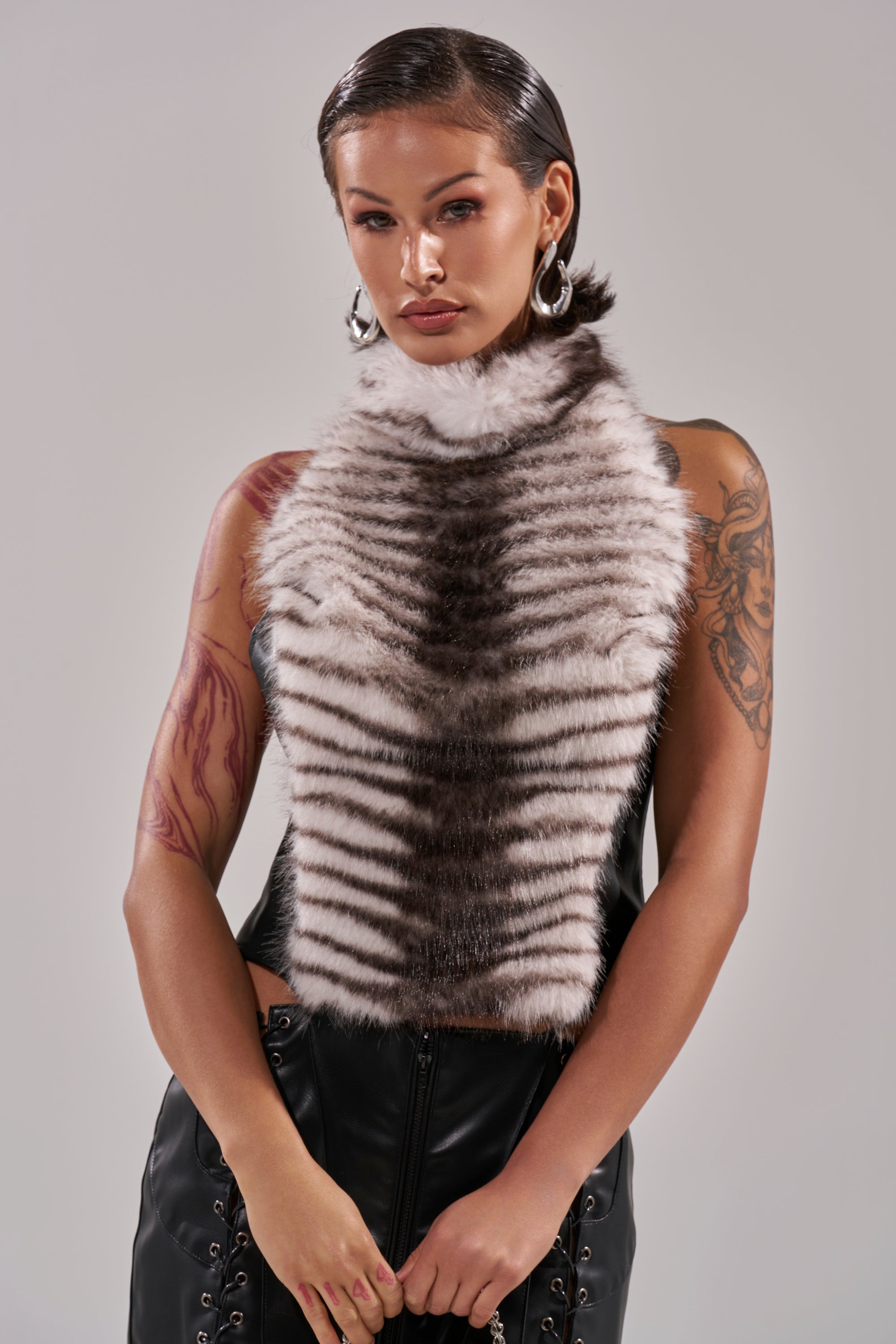 A woman with slicked-back hair, hoop earrings, and visible arm tattoos wears the I'M MY OWN SPIRIT ANIMAL MOCK NECK FUR TOP with stripes and faux leather pants, standing against a plain gray background. 2