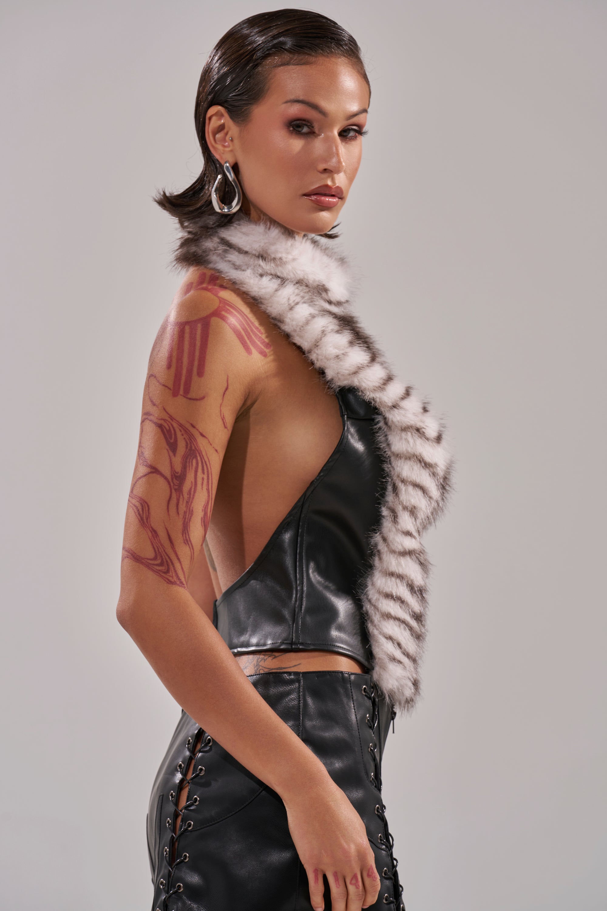 A woman with slicked-back dark hair and visible arm tattoos wears the I'M MY OWN SPIRIT ANIMAL MOCK NECK FUR TOP, paired with lace-up leather pants and hoop earrings, standing against a plain light background. 3
