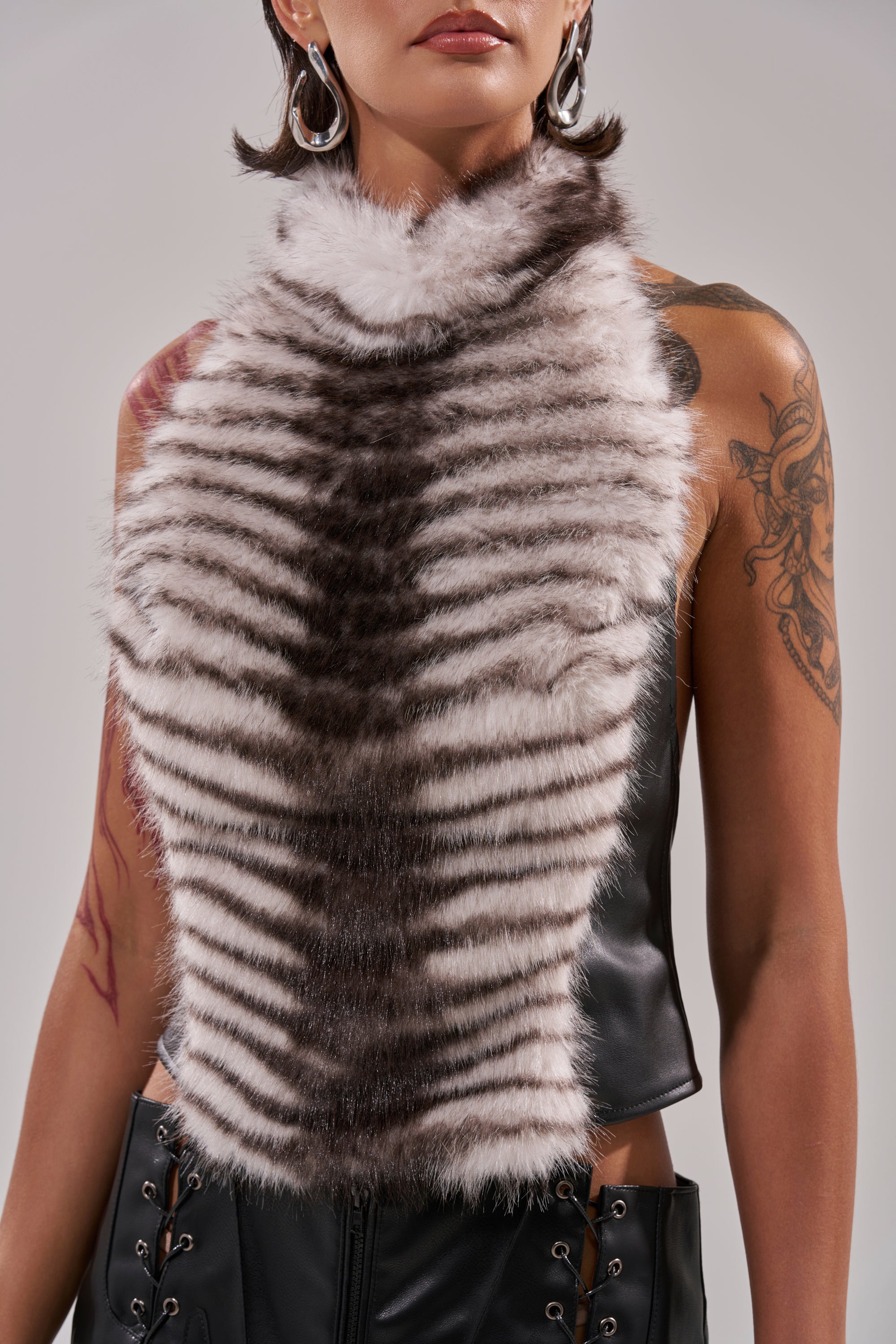A person wears the I'M MY OWN SPIRIT ANIMAL MOCK NECK FUR TOP, a sleeveless faux leather piece with a striped front. Hoop earrings and arm tattoos add edge. The photo is cropped at the shoulders and waist for a modern effect. 8