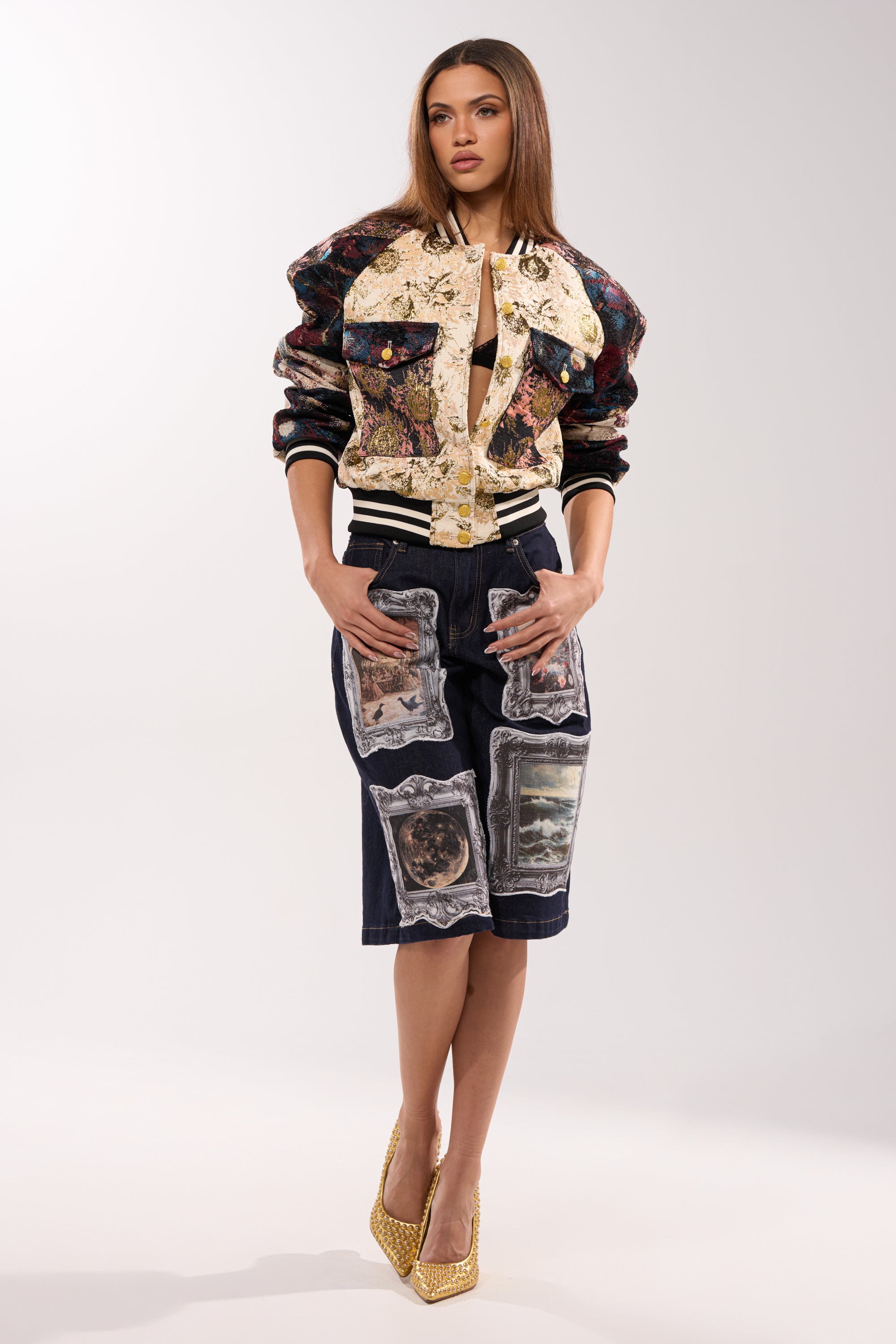 A woman stands confidently in the EGO CHECK PATCHWORK BOMBER over a denim skirt with an art-style patchwork pattern and yellow heels, posing with hands in her pockets against a plain white background.