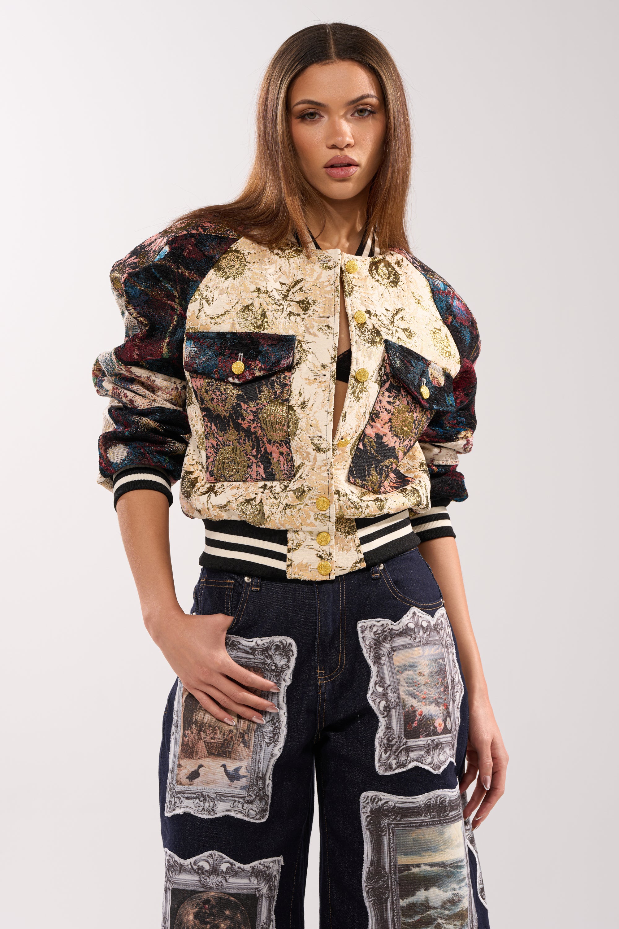 A woman with long straight hair wears the EGO CHECK PATCHWORK BOMBER and dark jeans with framed artwork prints, standing against a plain light background.