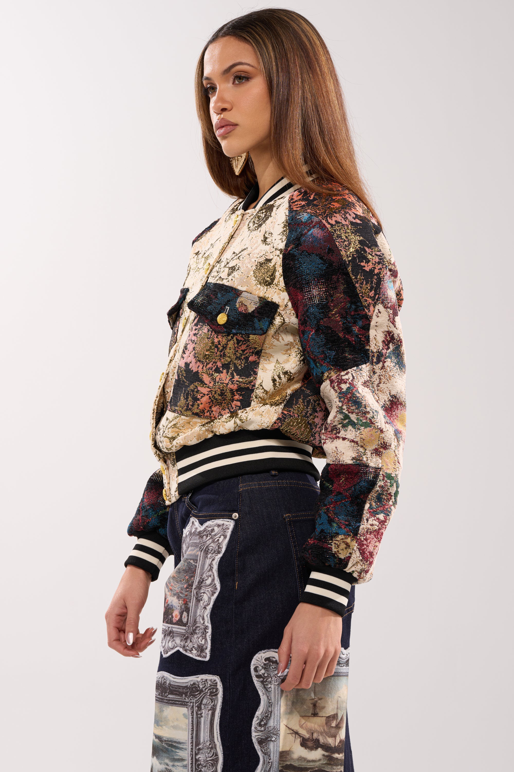 A woman stands in profile wearing the EGO CHECK PATCHWORK BOMBER with black-and-white striped cuffs and multicolored floral designs, paired with dark jeans featuring picture frame prints.