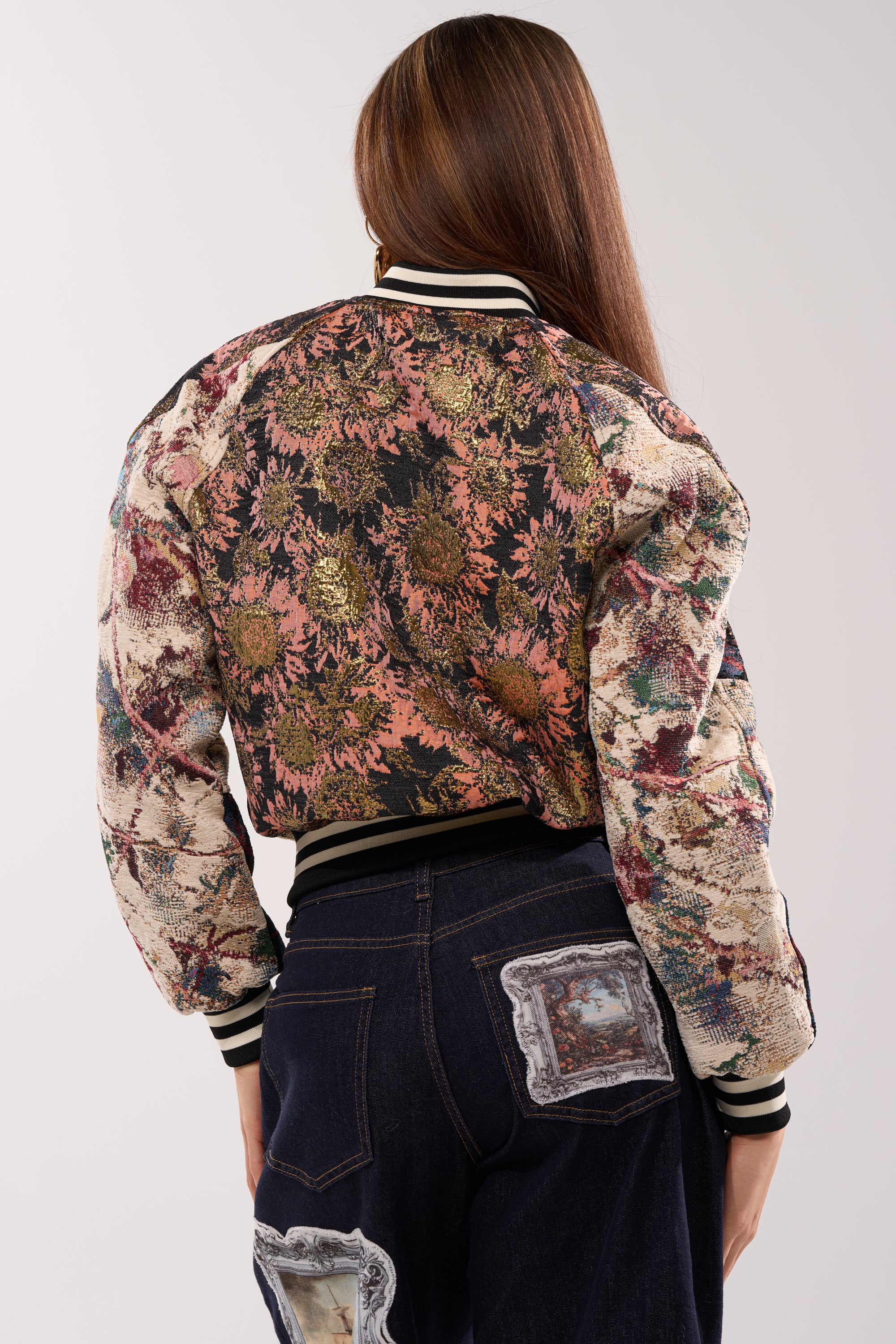A person with long brown hair is shown from the back, wearing the EGO CHECK PATCHWORK BOMBER and dark jeans with rectangular fabric patches against a plain, light-colored background.