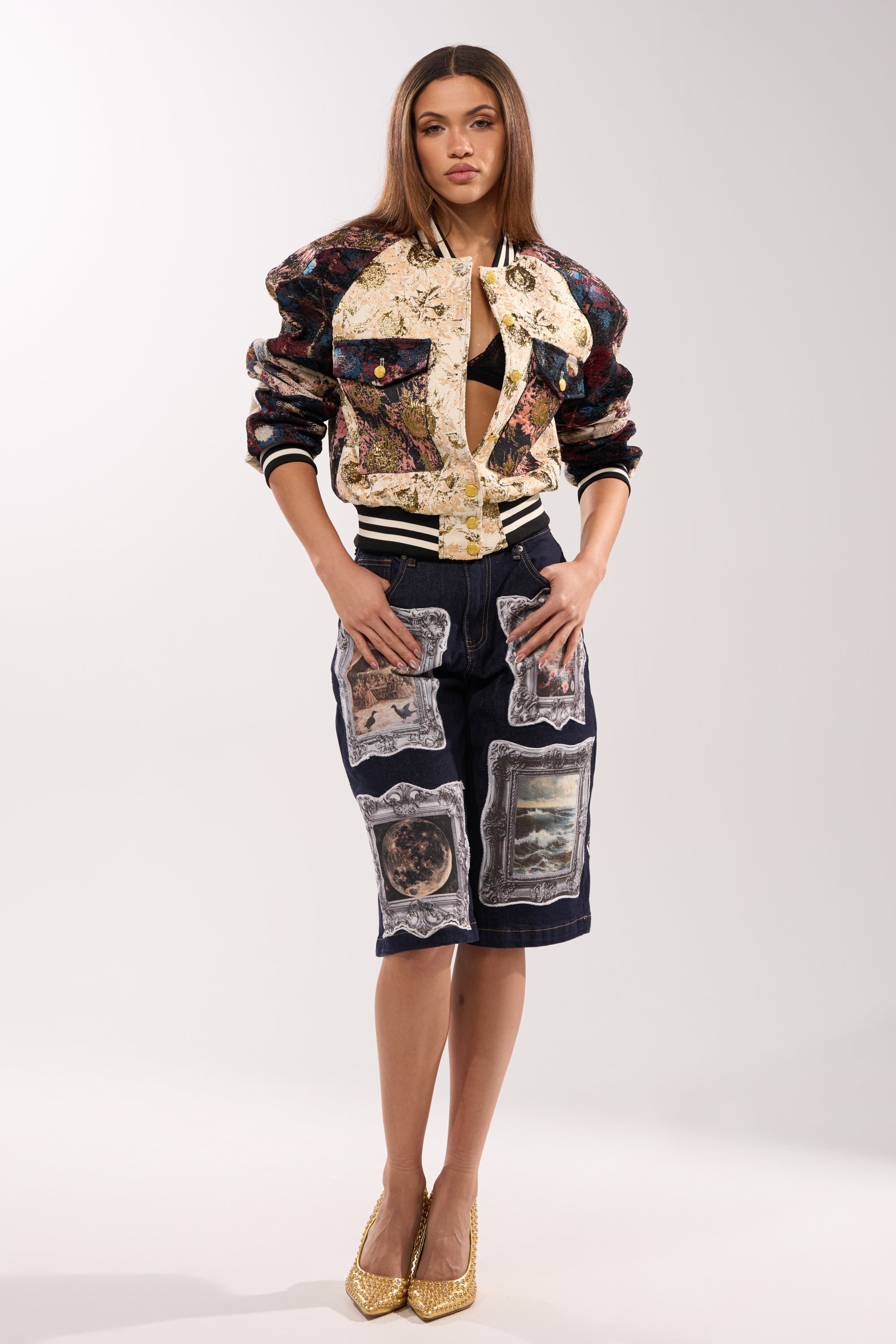 A woman models the EGO CHECK PATCHWORK BOMBER over a black top and knee-length dark shorts with scenic patches, paired with gold high heels against a plain light background.