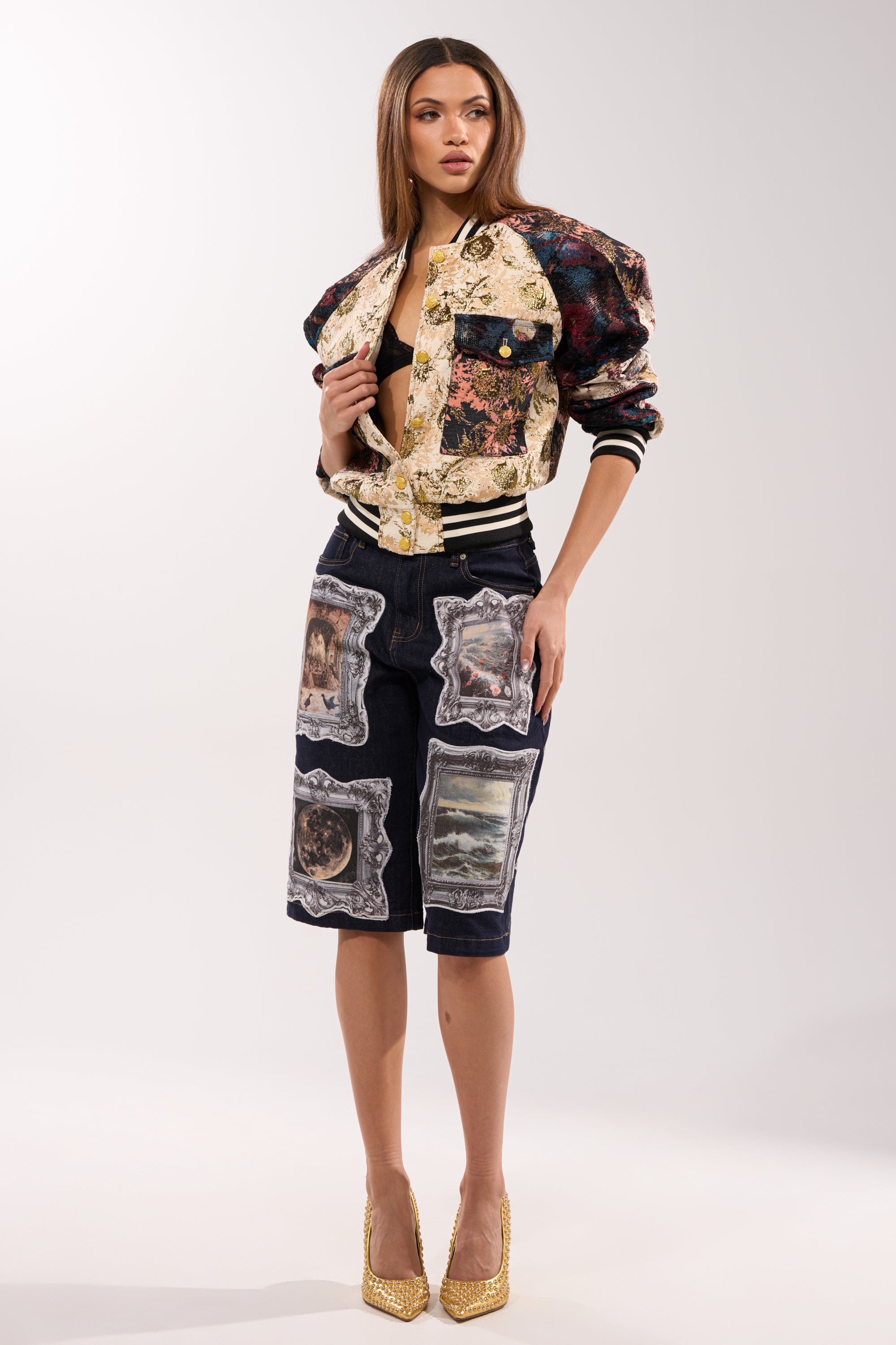 A woman poses in the EGO CHECK PATCHWORK BOMBER over a black cropped top and navy, art-print knee-length shorts, paired with gold heels, set against a plain light background.