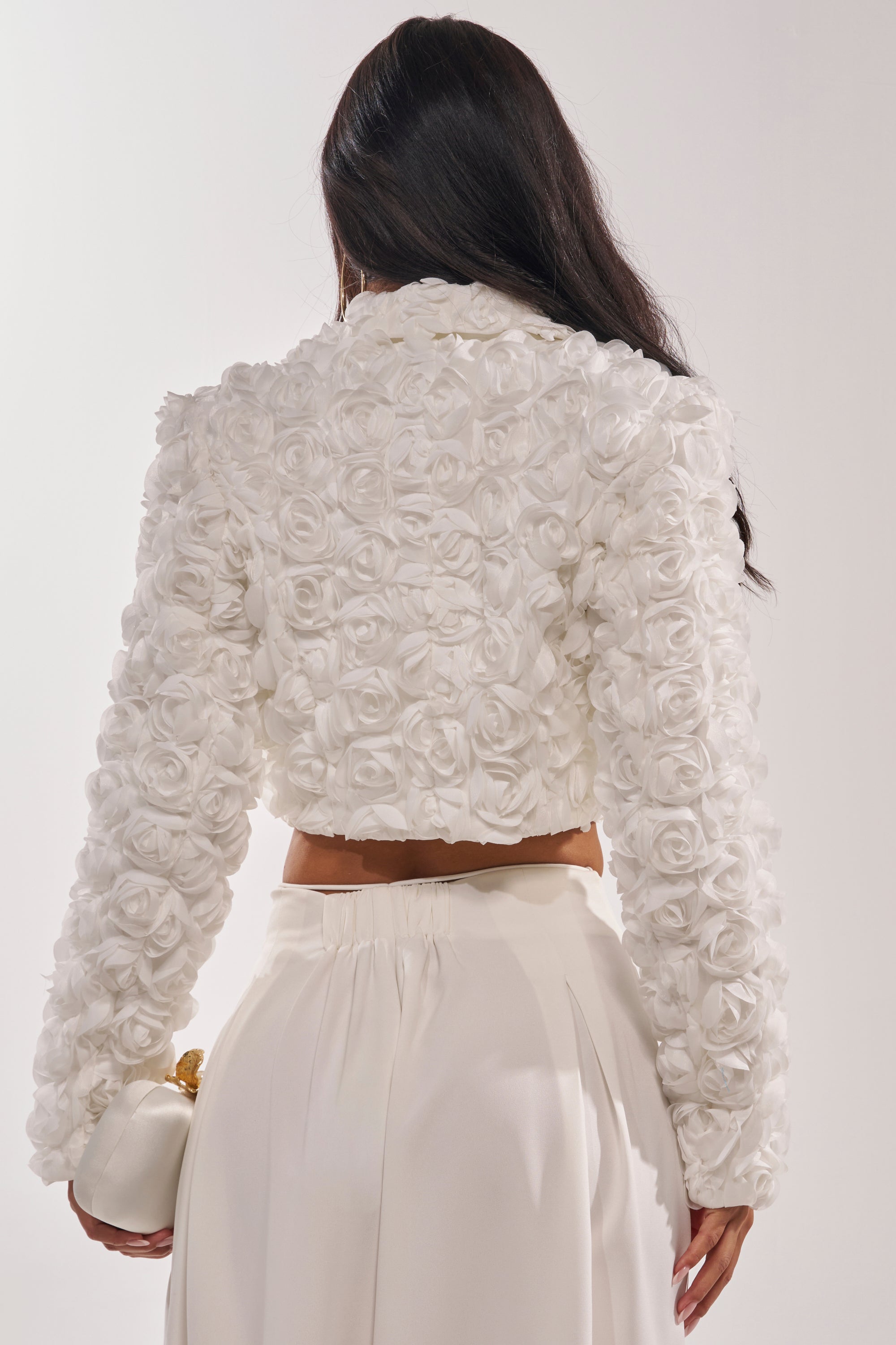 A woman with long dark hair is seen from the back, wearing the FAYE FLORAL APPLIQUE BLAZER—a white 3D rose cropped bridal blazer—paired with a high-waisted white skirt and holding a small white purse with a gold clasp. 7