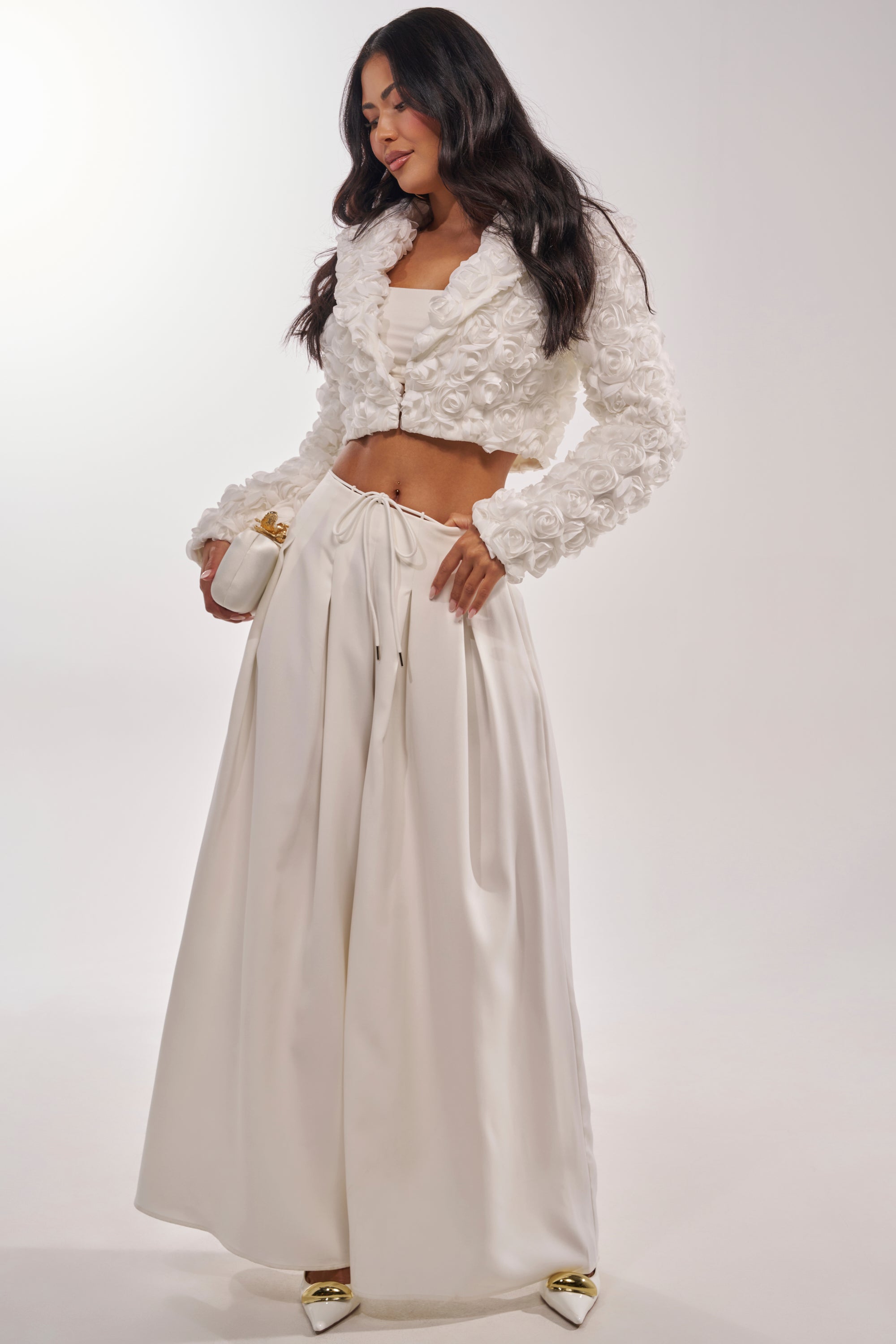 A woman poses in a chic all-white outfit featuring the FAYE FLORAL APPLIQUE BLAZER, matching top, wide-legged high-waisted pants, gold pointed heels, and a white clutch against a plain background. 8