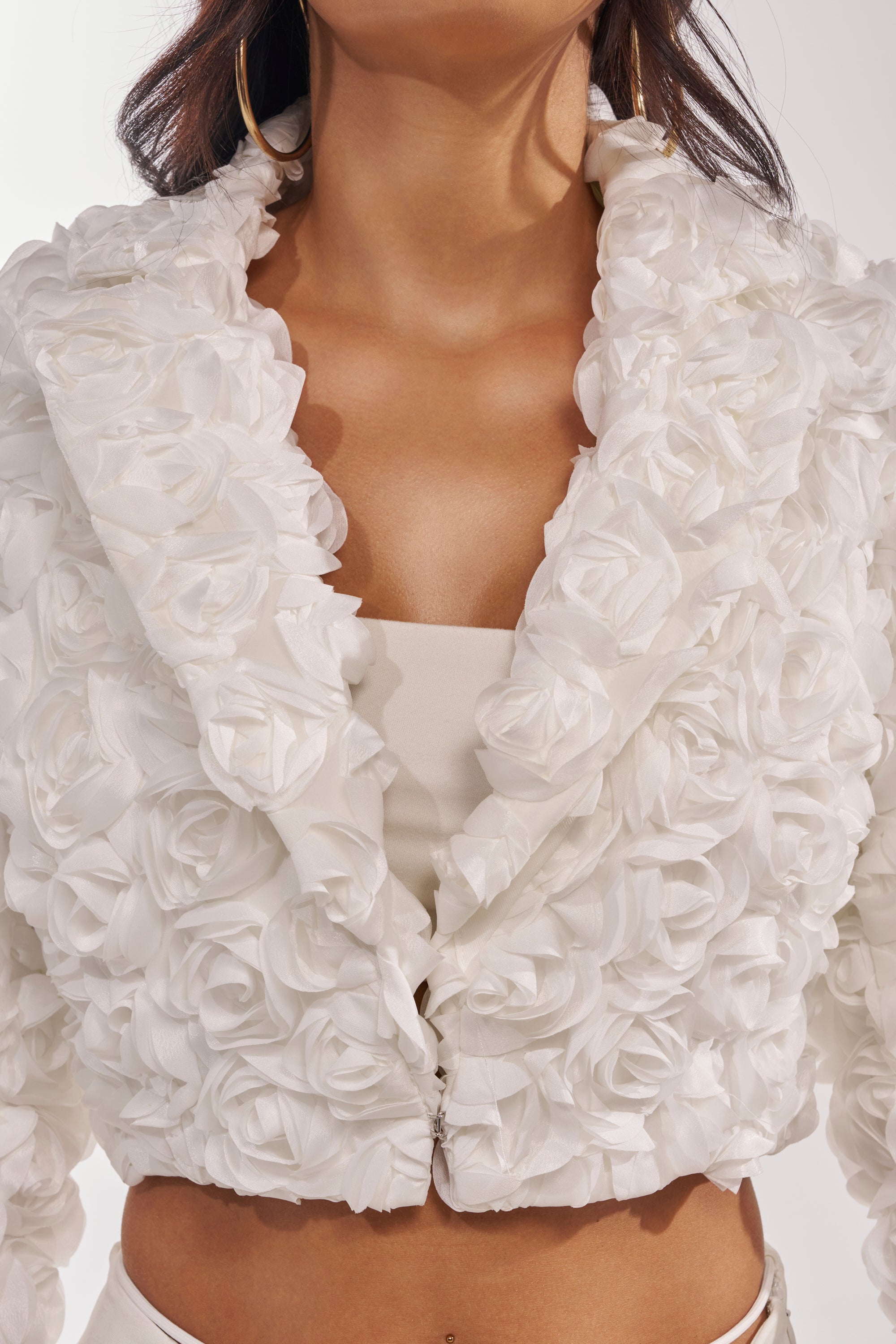 A woman wears the FAYE FLORAL APPLIQUE BLAZER, a white cropped jacket with fabric rosettes, over a matching top. The deep collar and sculptural floral design are visible, along with her torso and lower face. 11
