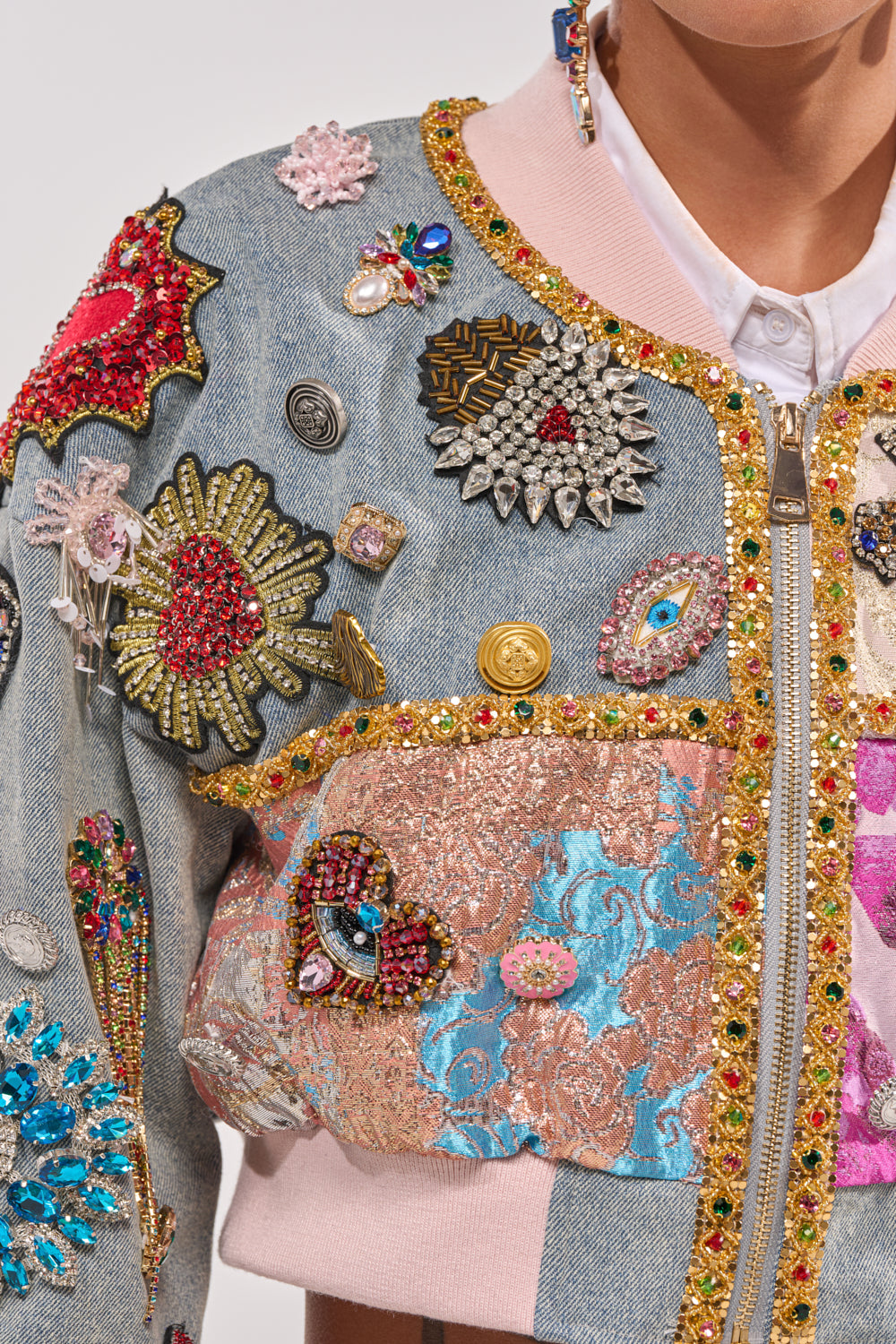 A close-up of a person wearing the IF YOU'RE DOWN EMBELLISHED DENIM BOMBER, adorned with colorful beaded, jeweled, and embroidered patches—flowers, eyes, abstract shapes—as well as a rhinestone brooch, pink collar, and shiny gold trim. 12