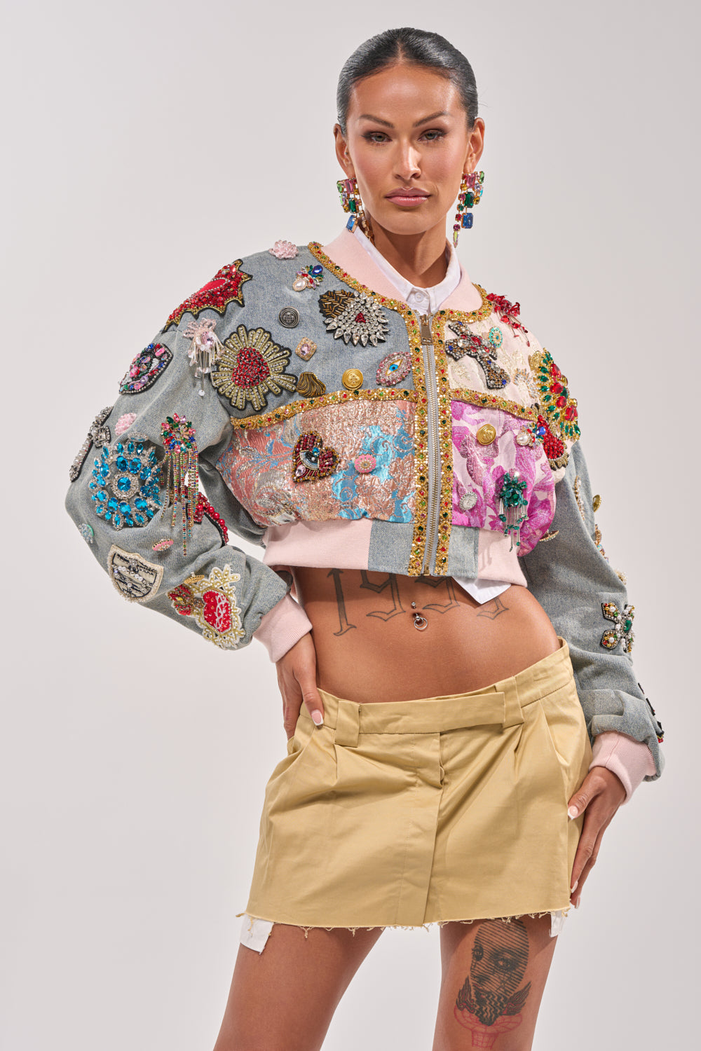 A woman with tattoos and slicked-back hair poses confidently in the IF YOU'RE DOWN EMBELLISHED DENIM BOMBER, bold earrings gleaming as she stands against a plain, light background. 1