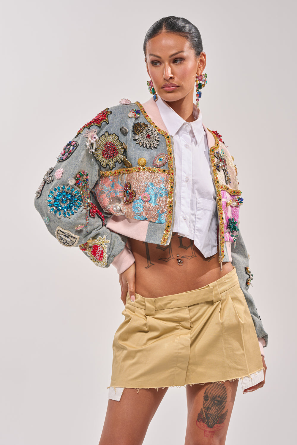 A woman poses confidently in the IF YOU'RE DOWN EMBELLISHED DENIM BOMBER, styled over a white shirt and beige pleated mini skirt, with statement earrings and a thigh tattoo on display. 2