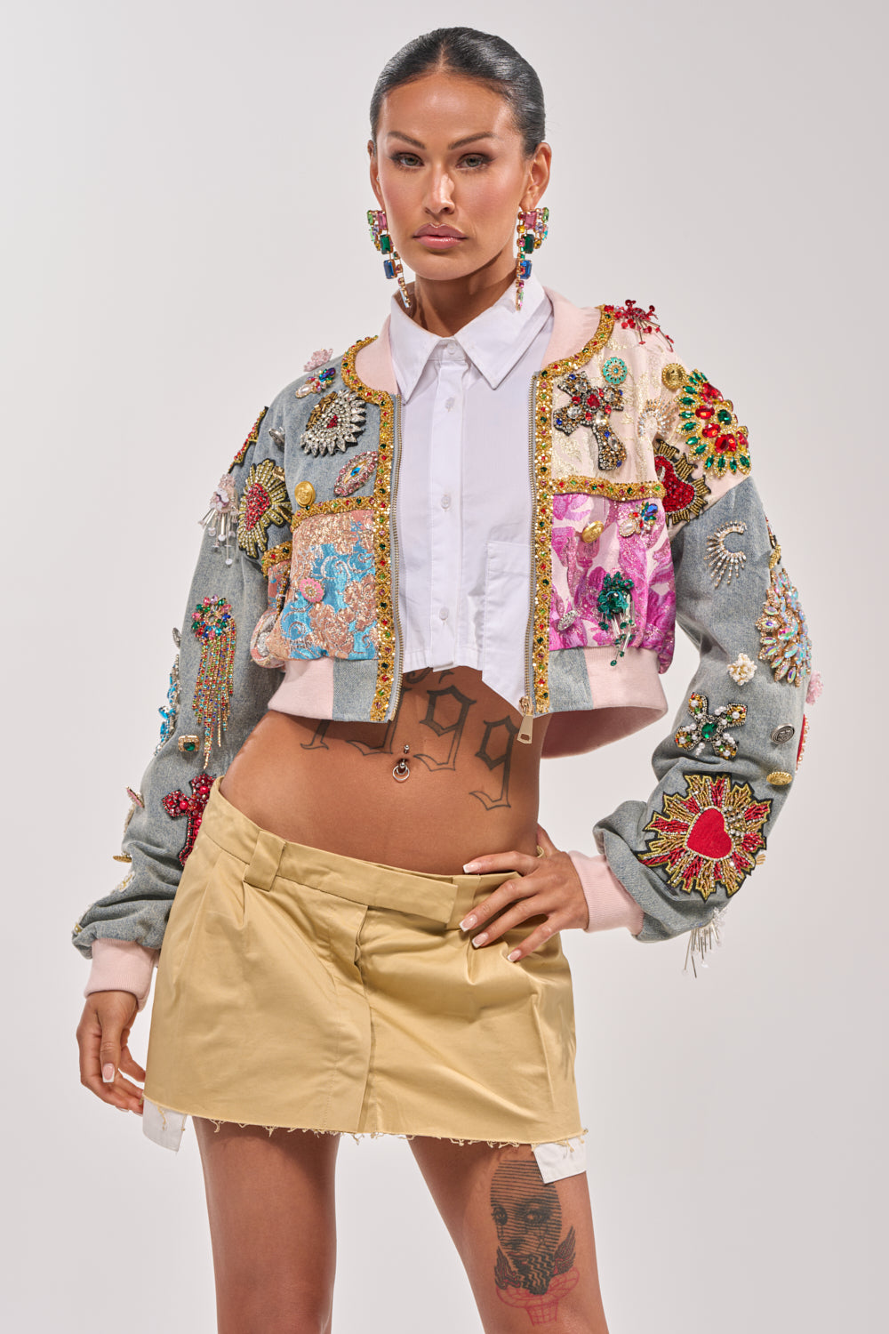 A person poses confidently in the IF YOU'RE DOWN EMBELLISHED DENIM BOMBER over a white collared shirt, paired with a short beige skirt and vibrant earrings, showcasing tattoos on their midriff and thigh. 4