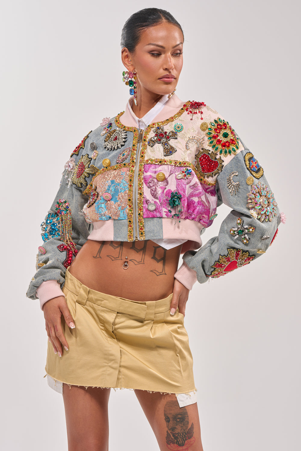 A woman wearing the "IF YOU'RE DOWN EMBELLISHED DENIM BOMBER" and a beige mini skirt stands with one hand on her hip, showcasing torso and thigh tattoos. Her hair is tied back against a plain light gray background. 5