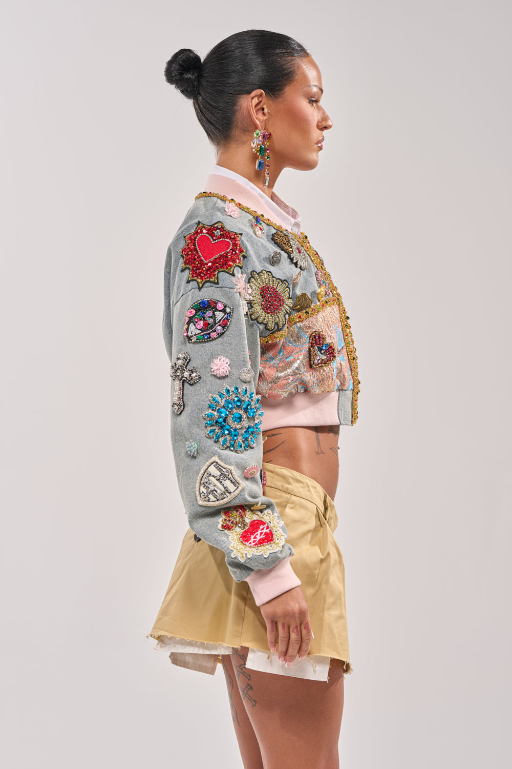A woman in profile with a sleek bun stands against a plain background, wearing the IF YOU'RE DOWN EMBELLISHED DENIM BOMBER, featuring colorful patches and a pink collar, paired with a beige pleated mini skirt and beaded earrings. 6
