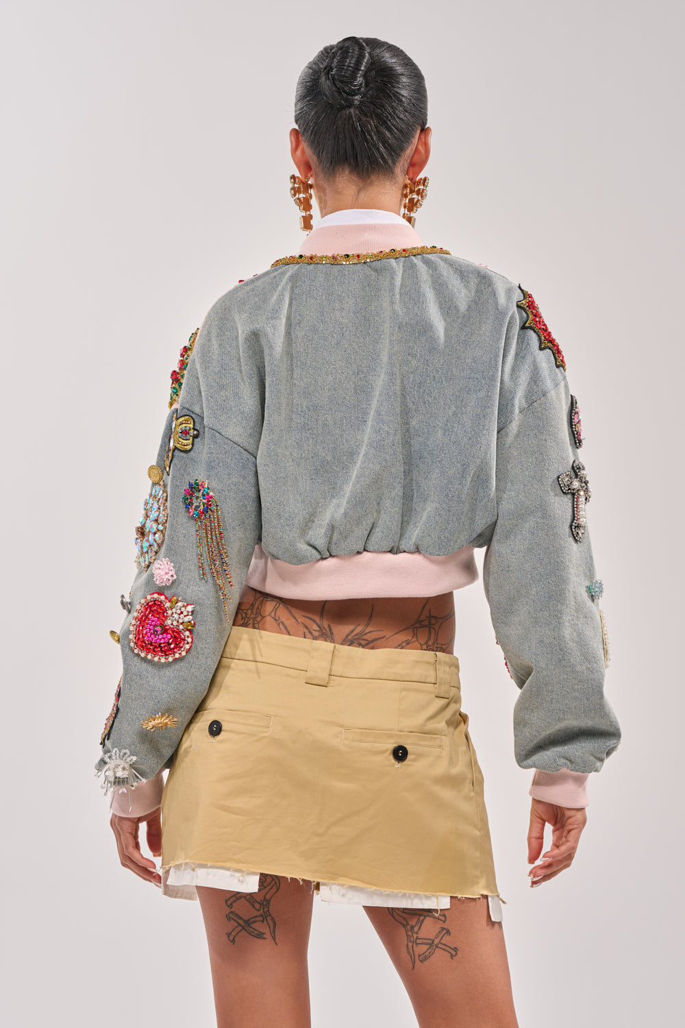 A person faces away, wearing the IF YOU'RE DOWN EMBELLISHED DENIM BOMBER with colorful rhinestone patches on the sleeves, paired with a tan mini skirt. Their hair is slicked back and lower back tattoos are visible. 7