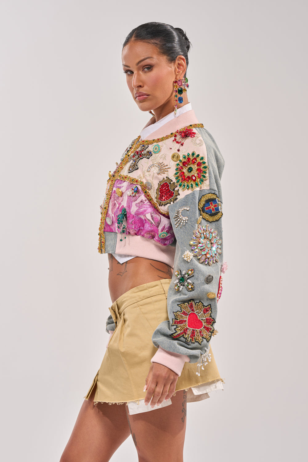 A woman in profile wears the IF YOU'RE DOWN EMBELLISHED DENIM BOMBER with jewels and patches, paired with a tan mini skirt. Her hair is tied back and she accessorizes with bold, colorful earrings. 8