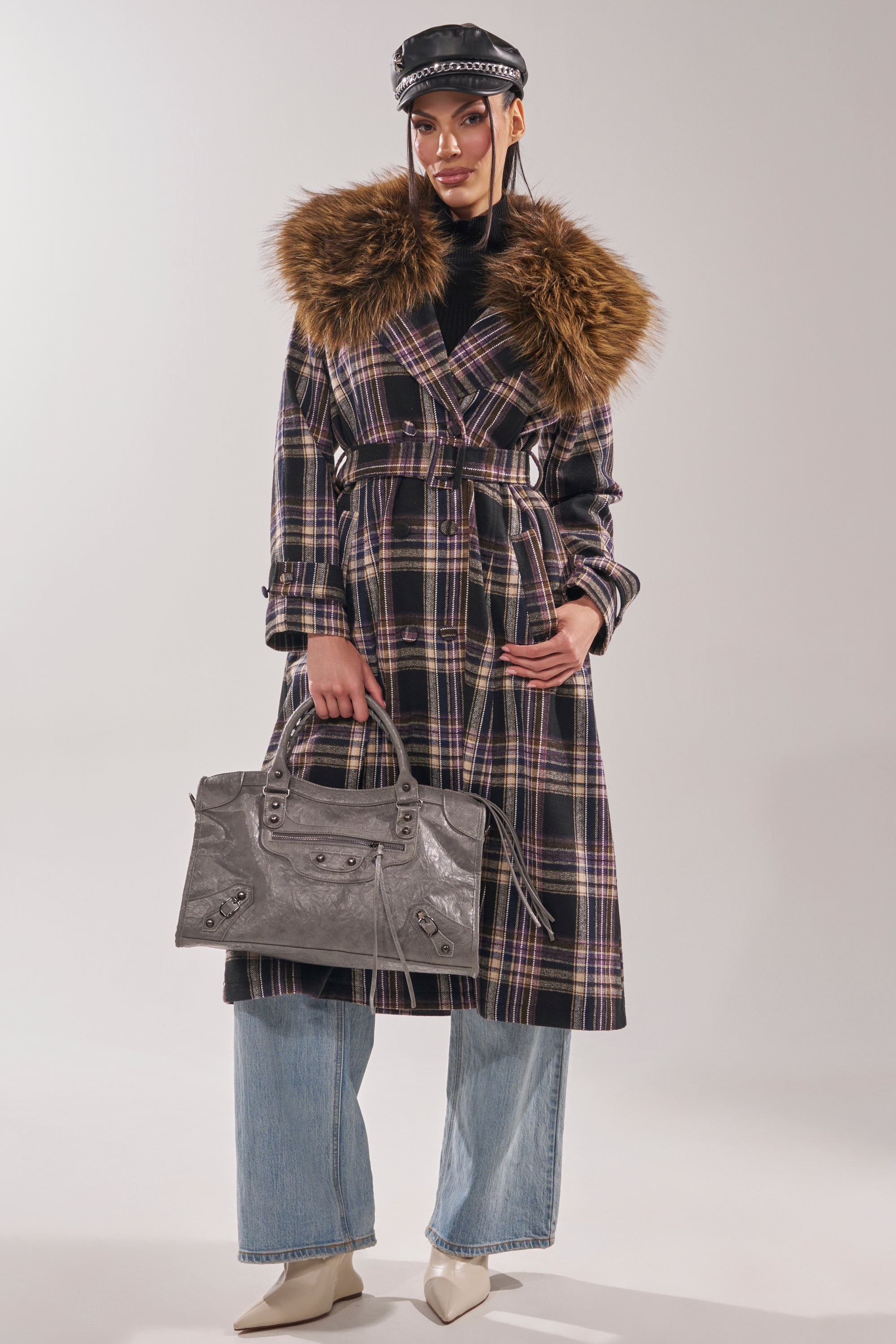 A person wears the SWEPT UP FUR TRIM TRENCH with a large faux fur collar, light blue wide-leg jeans, cream pointed boots, a black top, black leather cap, and carries a large gray handbag.