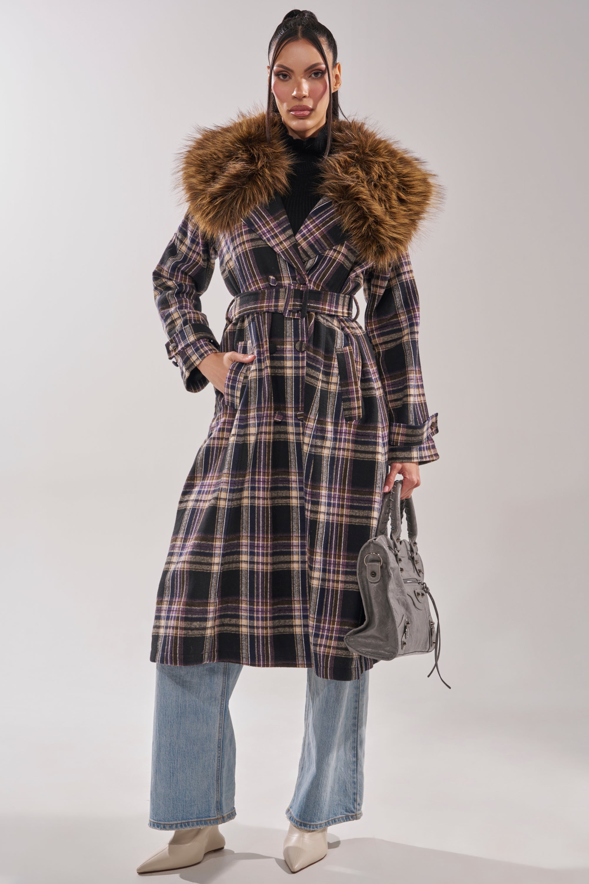 A woman confidently models the SWEPT UP FUR TRIM TRENCH, featuring a large faux fur collar, paired with wide-leg jeans, white pointed-toe boots, and a gray handbag against a plain background.