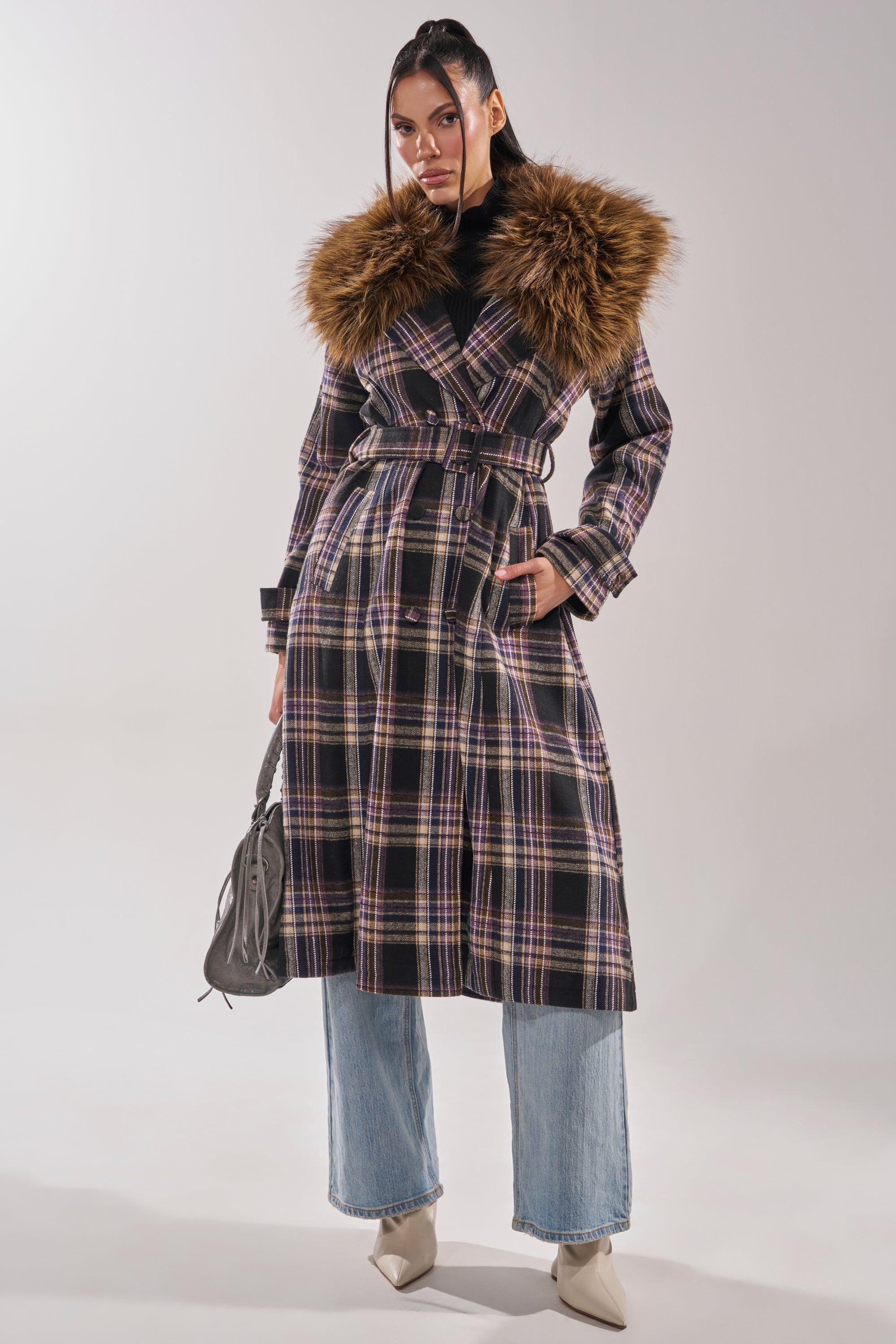 A person models the SWEPT UP FUR TRIM TRENCH, featuring a plaid design and wide faux fur collar, belted at the waist, paired with light blue jeans, pointed-toe shoes, and a gray handbag against a plain background.