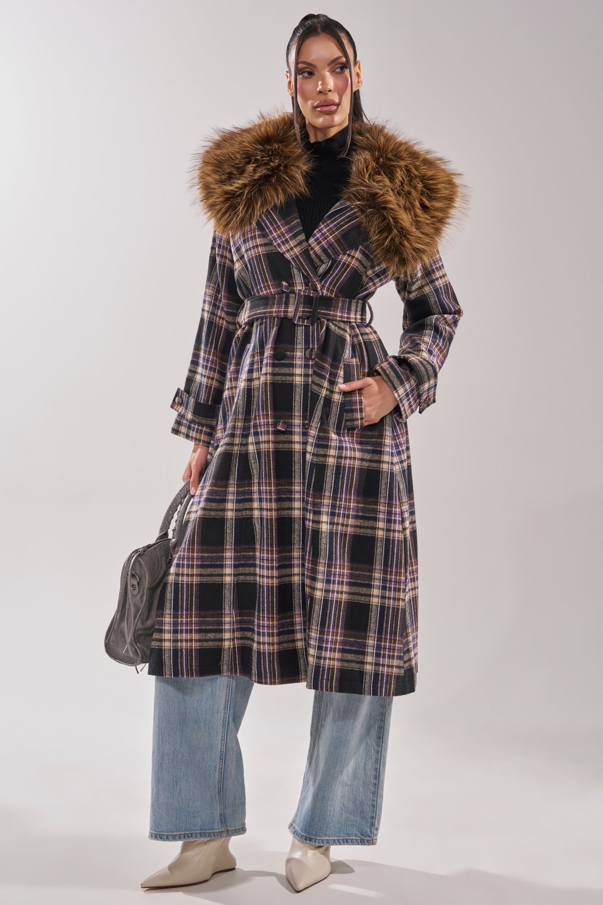 A woman models the SWEPT UP FUR TRIM TRENCH, a longline plaid coat with a bold faux fur collar, paired with light blue wide-leg jeans, white pointed shoes, and a gray handbag as she stands confidently against a plain background.