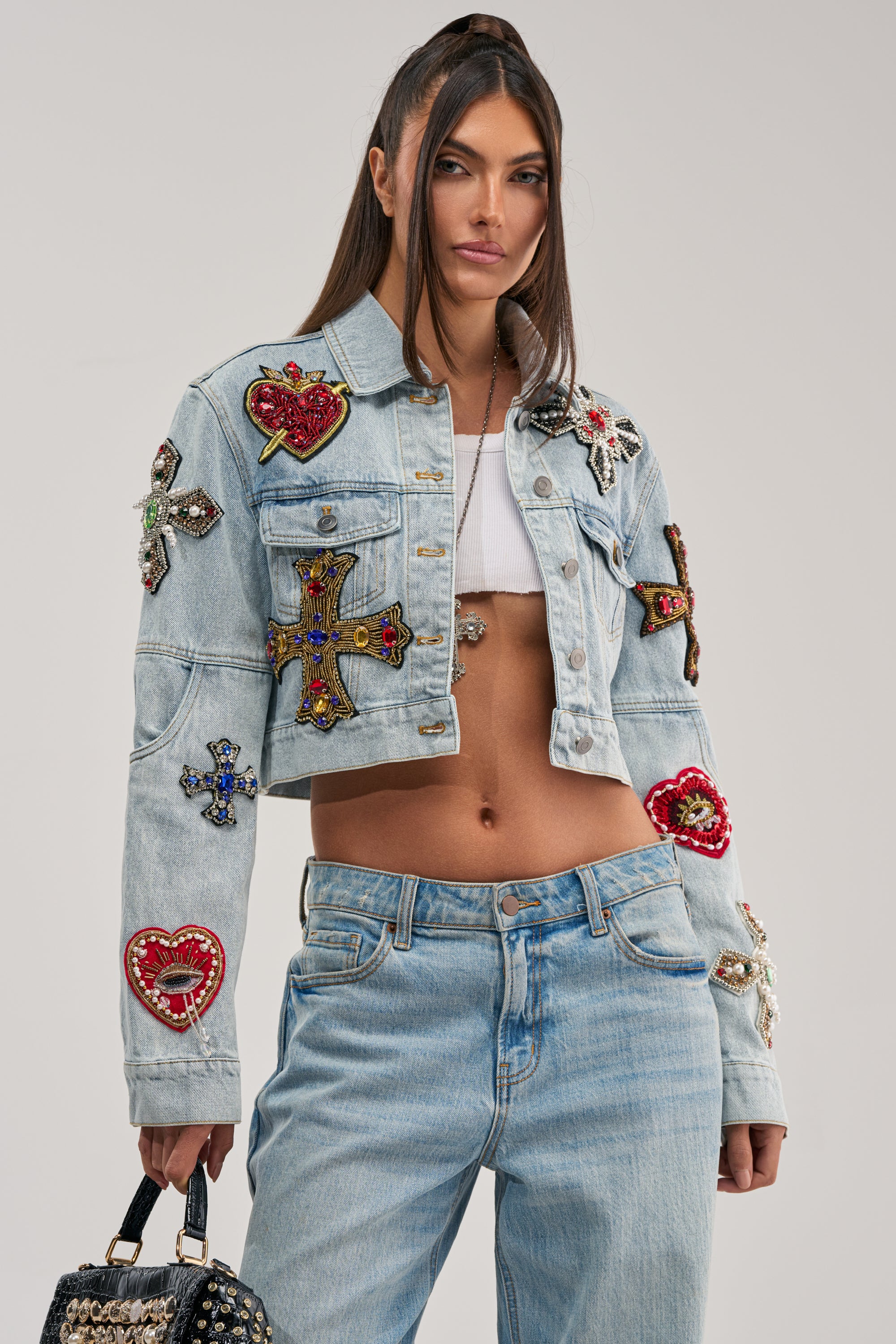 A woman wearing the GIVE AND TAKE EMBELLISHED DENIM JACKET with colorful bead patches and matching jeans holds a black studded handbag against a plain background.